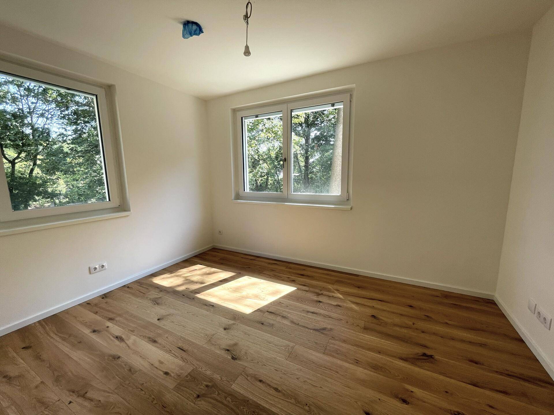 Helles Schlafzimmer mit Holzboden und zwei Fenstern, die einen schönen Blick ins Grüne bieten.