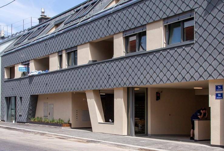Moderne Wohnhausfassade mit Schieferdach und integrierten Balkonen an einer Straße.