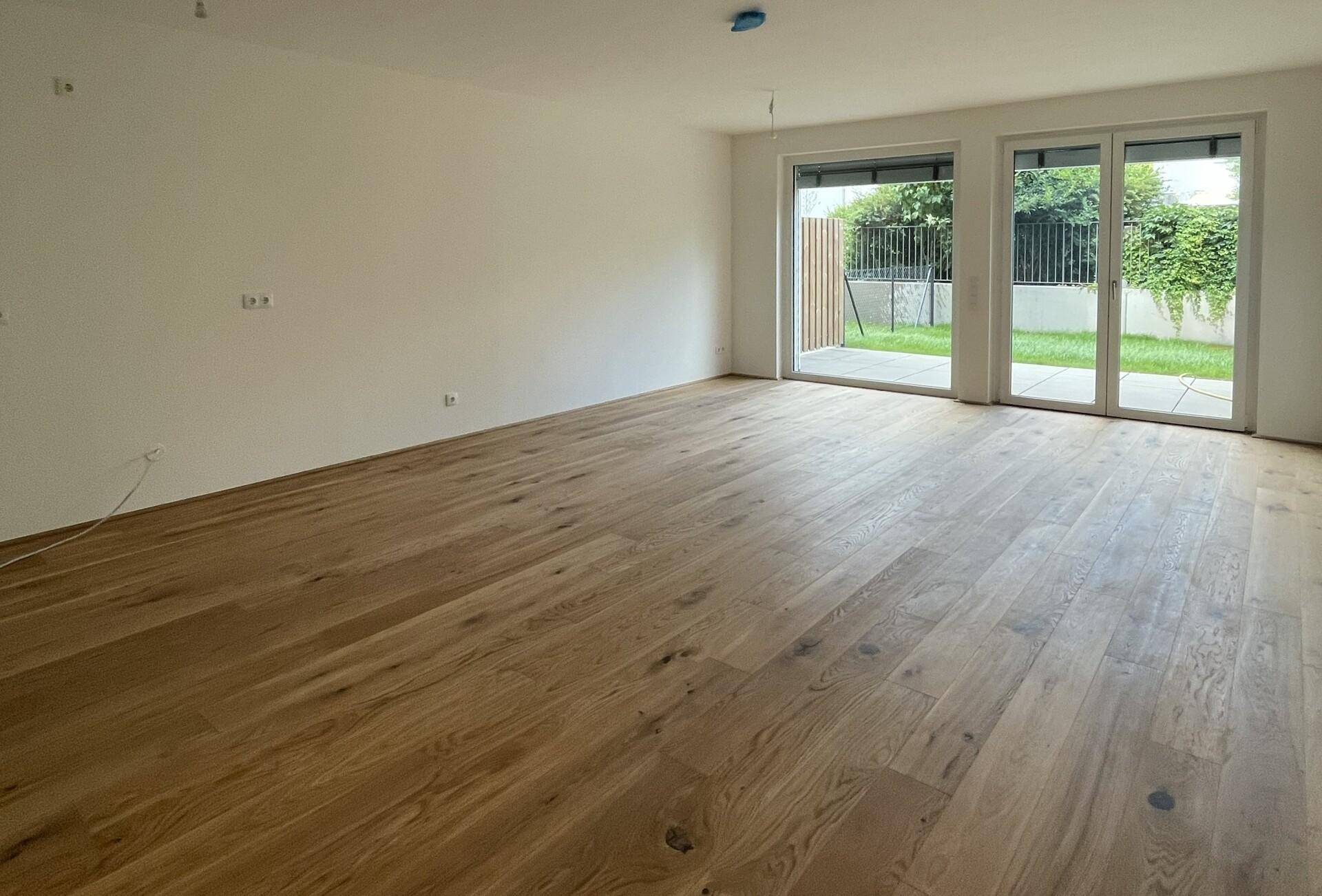 Geräumiges, leeres Wohnzimmer mit hellem Holzboden und großen Fenstern zum Garten.