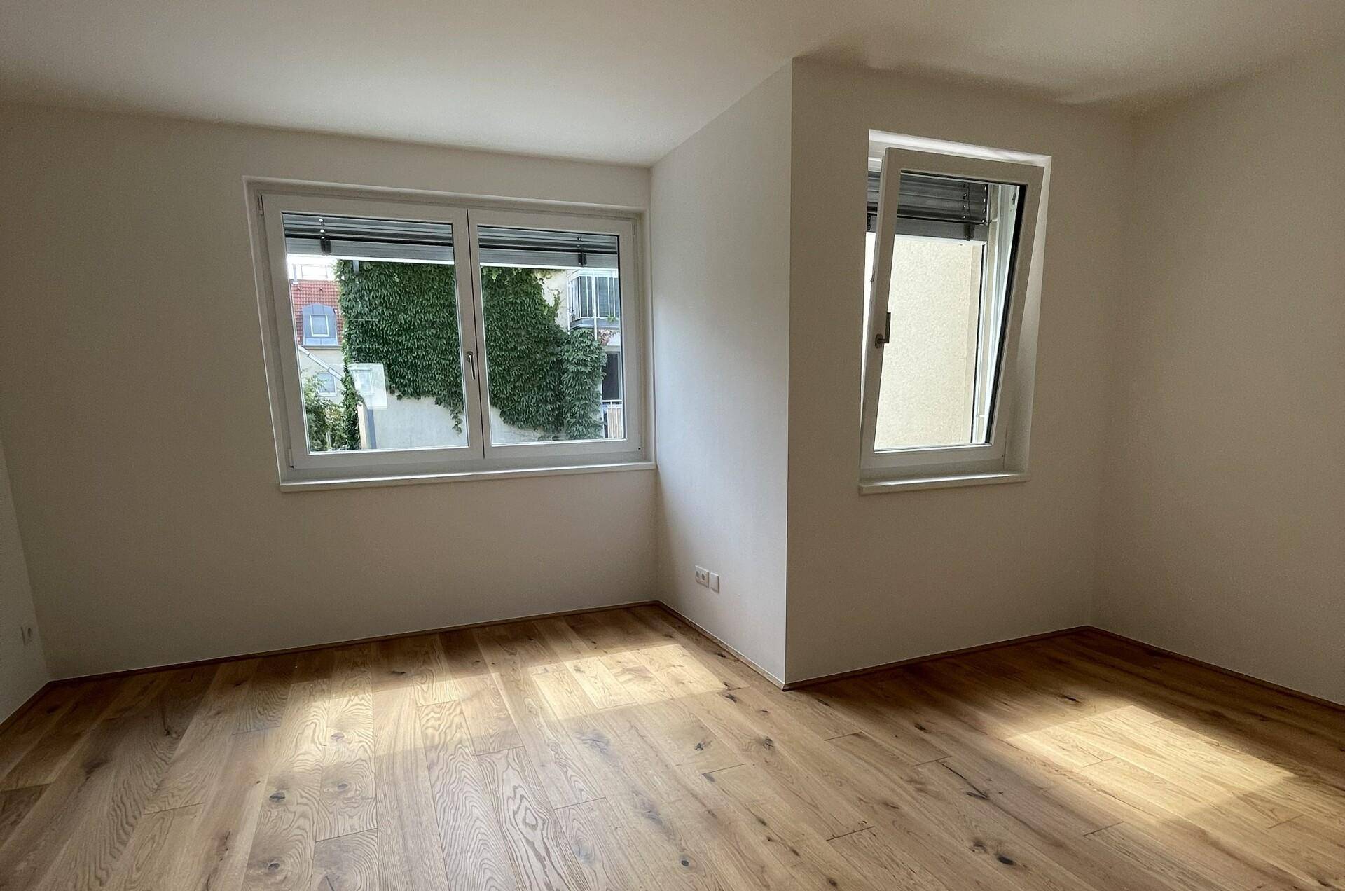 Helles Schlafzimmer mit Holzboden und zwei Fenstern, die viel Tageslicht hereinlassen.