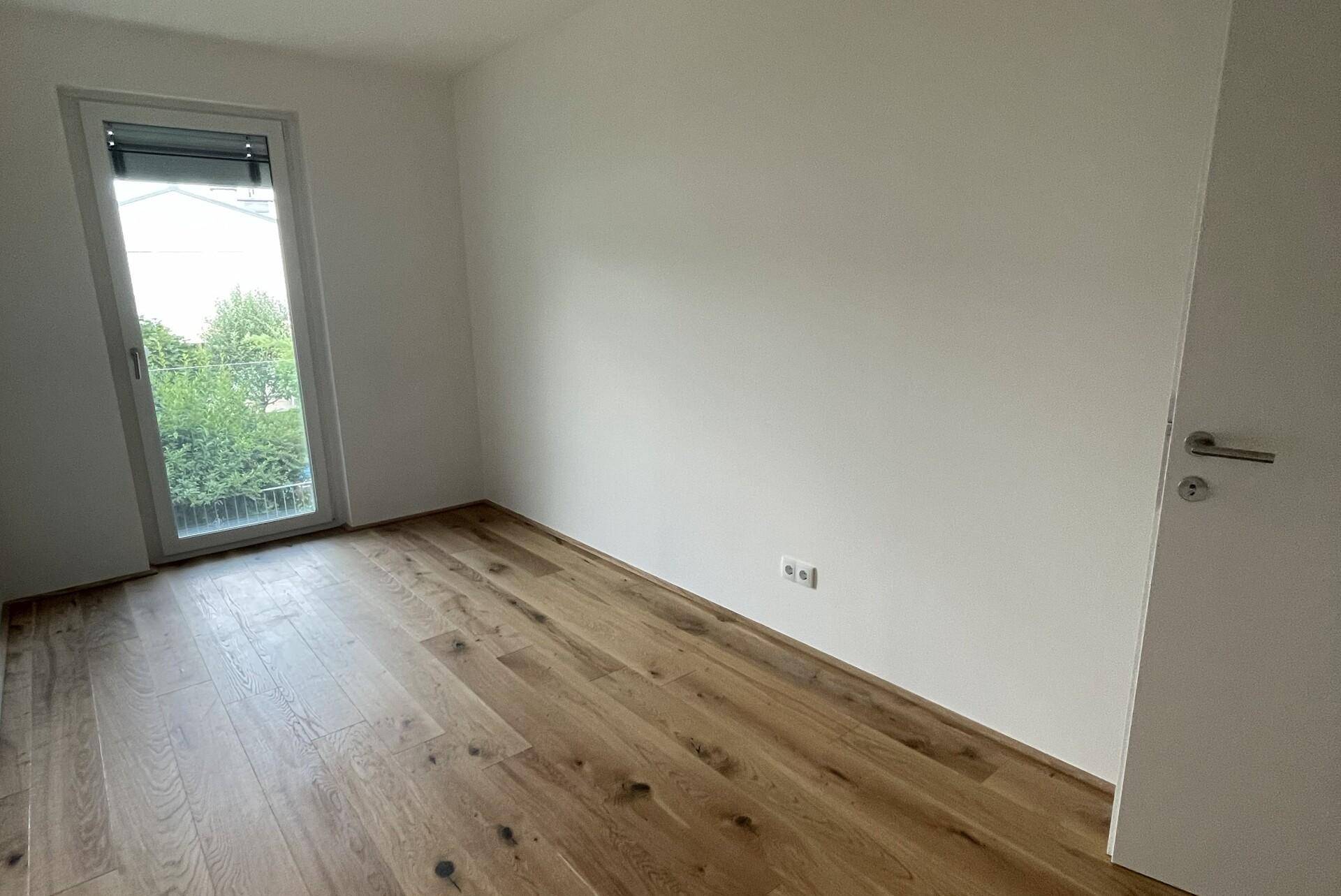Leeres Schlafzimmer mit Holzboden und einer Tür, die zu einem Balkon oder einer Terrasse führt.
