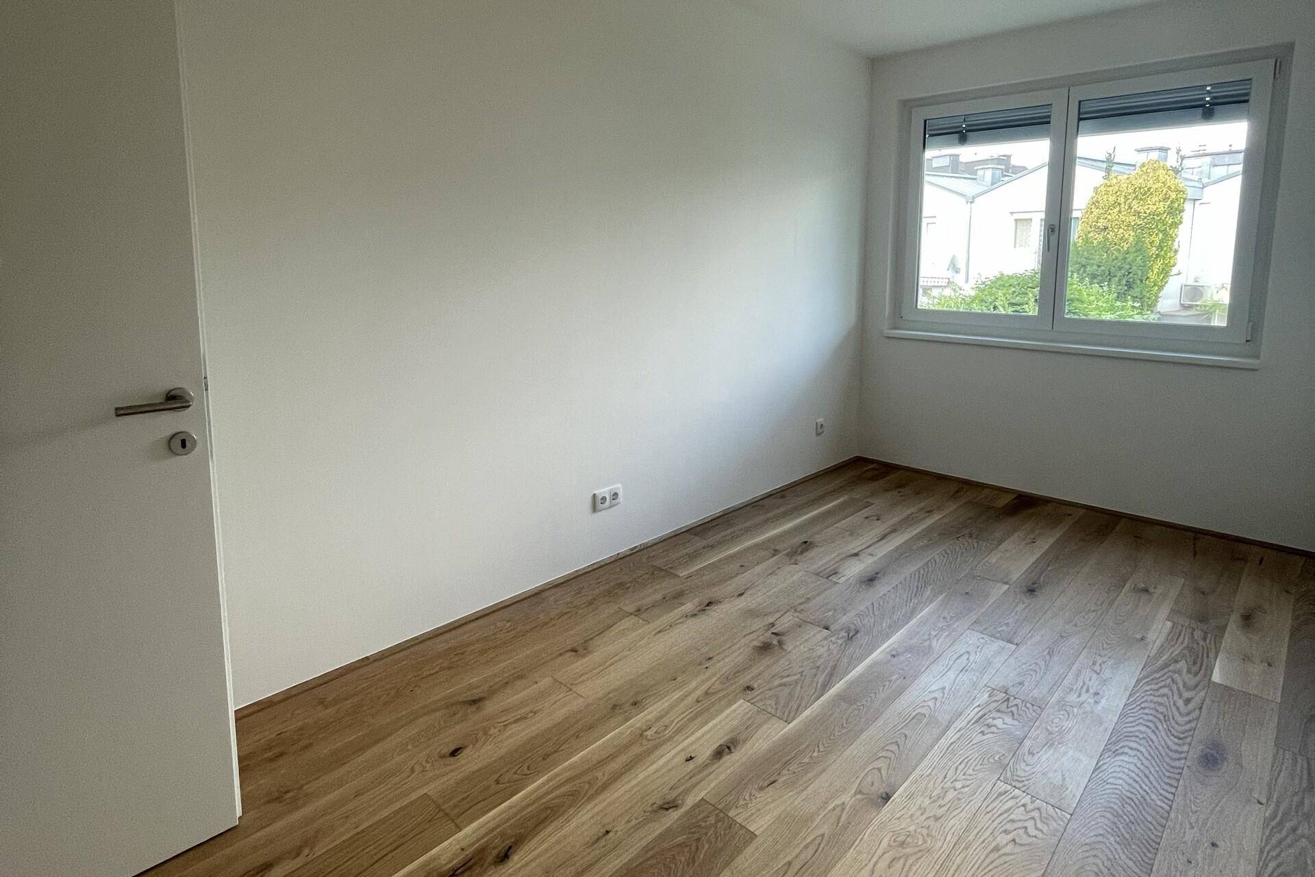 Einfaches Schlafzimmer mit Holzboden und einem Fenster, das den Blick auf die Nachbarschaft freigibt.