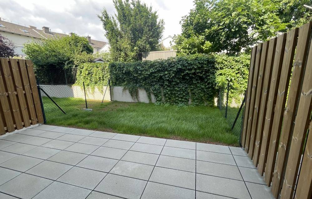 Private Terrasse mit gepflastertem Bereich und angrenzendem kleinen Garten, umzäunt von einem Holzzaun.