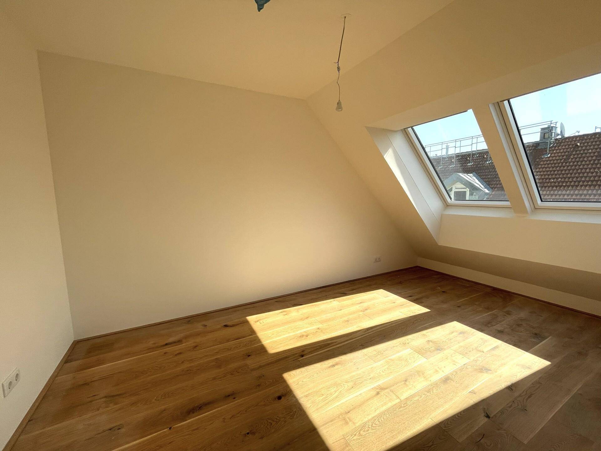 Sonniger Raum mit Holzdielenboden, weißen Wänden und zwei schrägen Dachfenstern, die viel Licht hereinlassen.