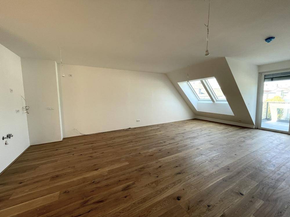 Heller, leerer Raum mit Holzdielenboden, weißen Wänden und großem Dachfenster mit Balkonzugang.