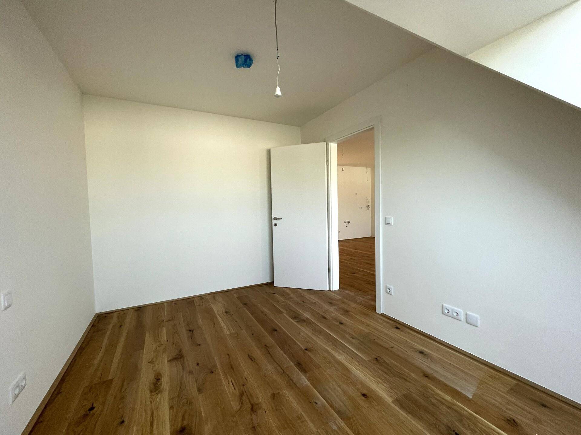 Leerer Raum mit Holzdielenboden und weiß gestrichenen Wänden, eine offene Tür führt in einen weiteren Raum.