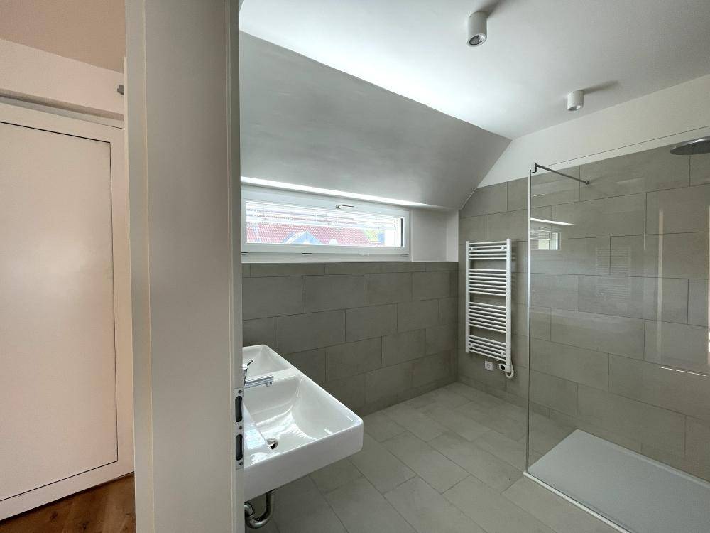 Modernes Badezimmer mit grauen Fliesen, ebenerdiger Dusche, Einzelwaschbecken und Fenster unter Schrägdach.