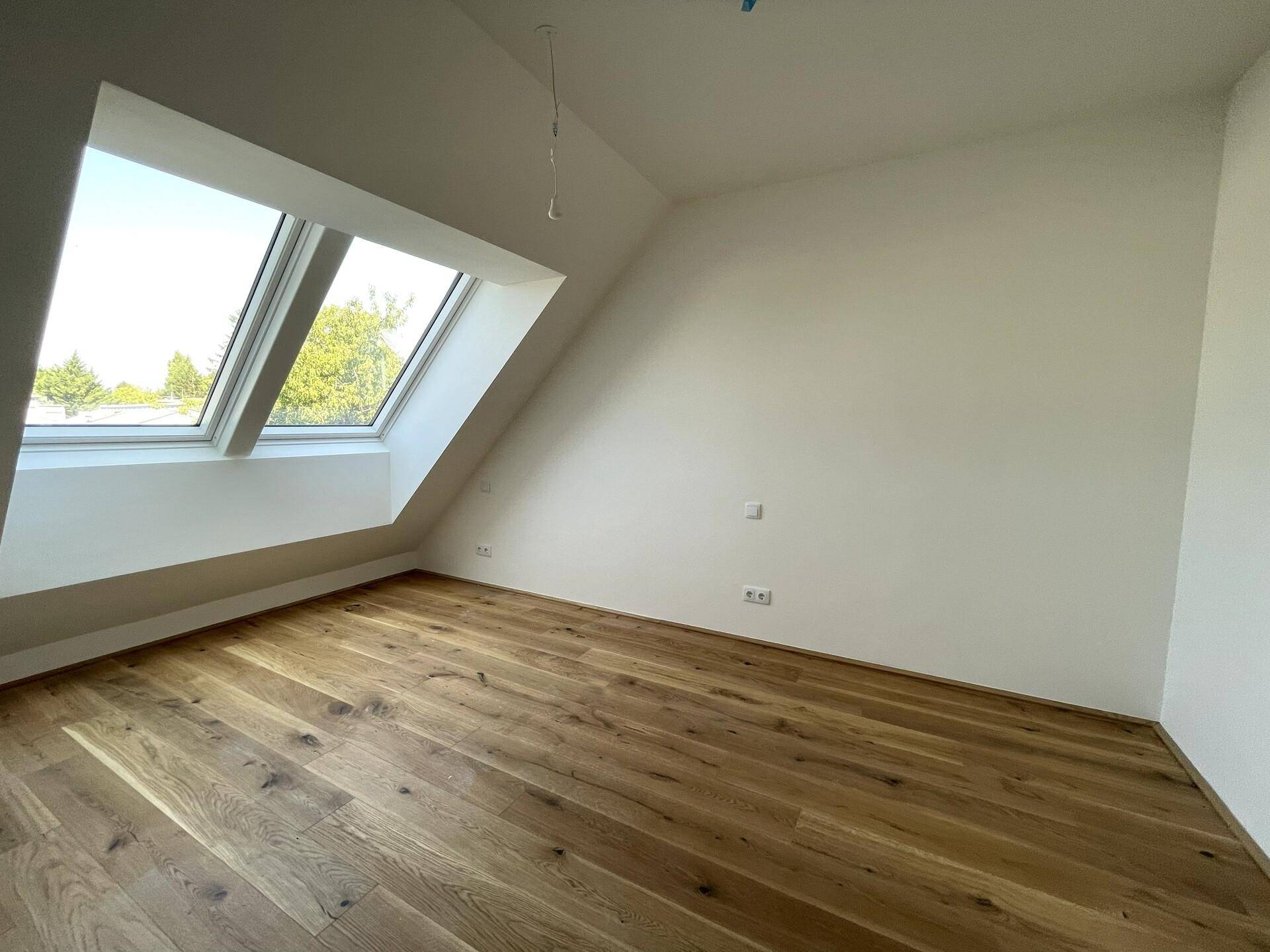 Heller Raum mit Holzdielenboden, weißen Wänden und Dachfenstern mit Blick ins Grüne.
