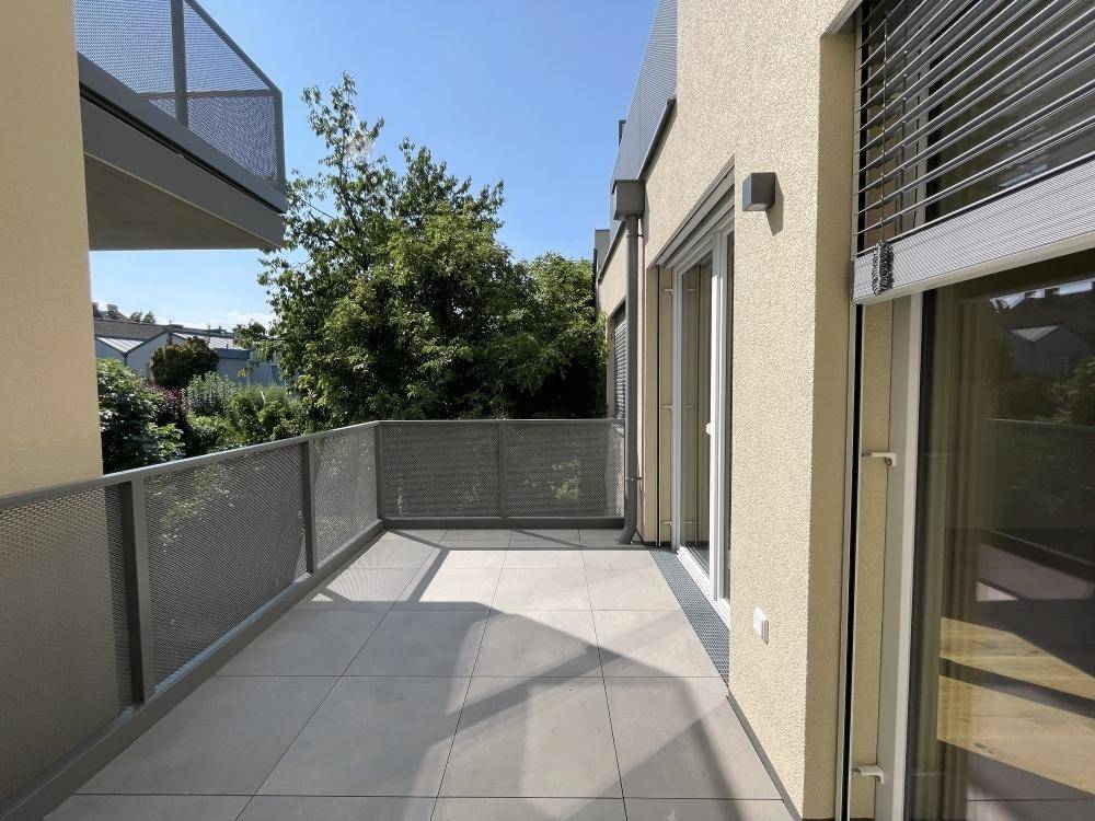 Heller Balkon mit modernem Metallgeländer und Fliesenboden, umgeben von Bäumen und Grünflächen.
