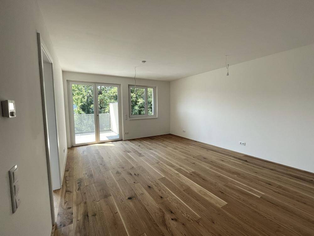 Helles, leeres Wohnzimmer mit neuem Holzparkettboden und Zugang zu einem Balkon.