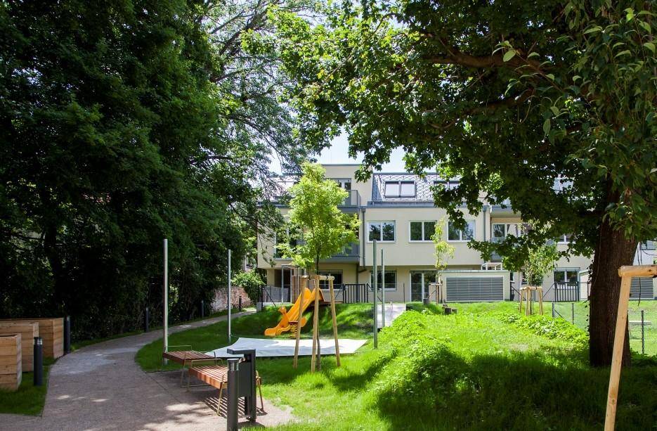 Grüner Gemeinschaftsgarten mit Spielplatz, Bänken und Bäumen vor einem Wohngebäude.