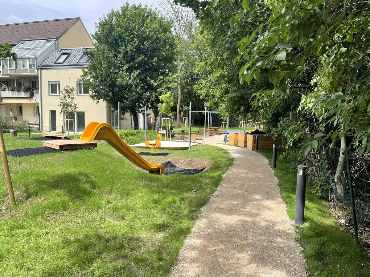 Grüner Weg führt zu einem Spielplatz mit Rutsche und Schaukeln, umgeben von Bäumen.