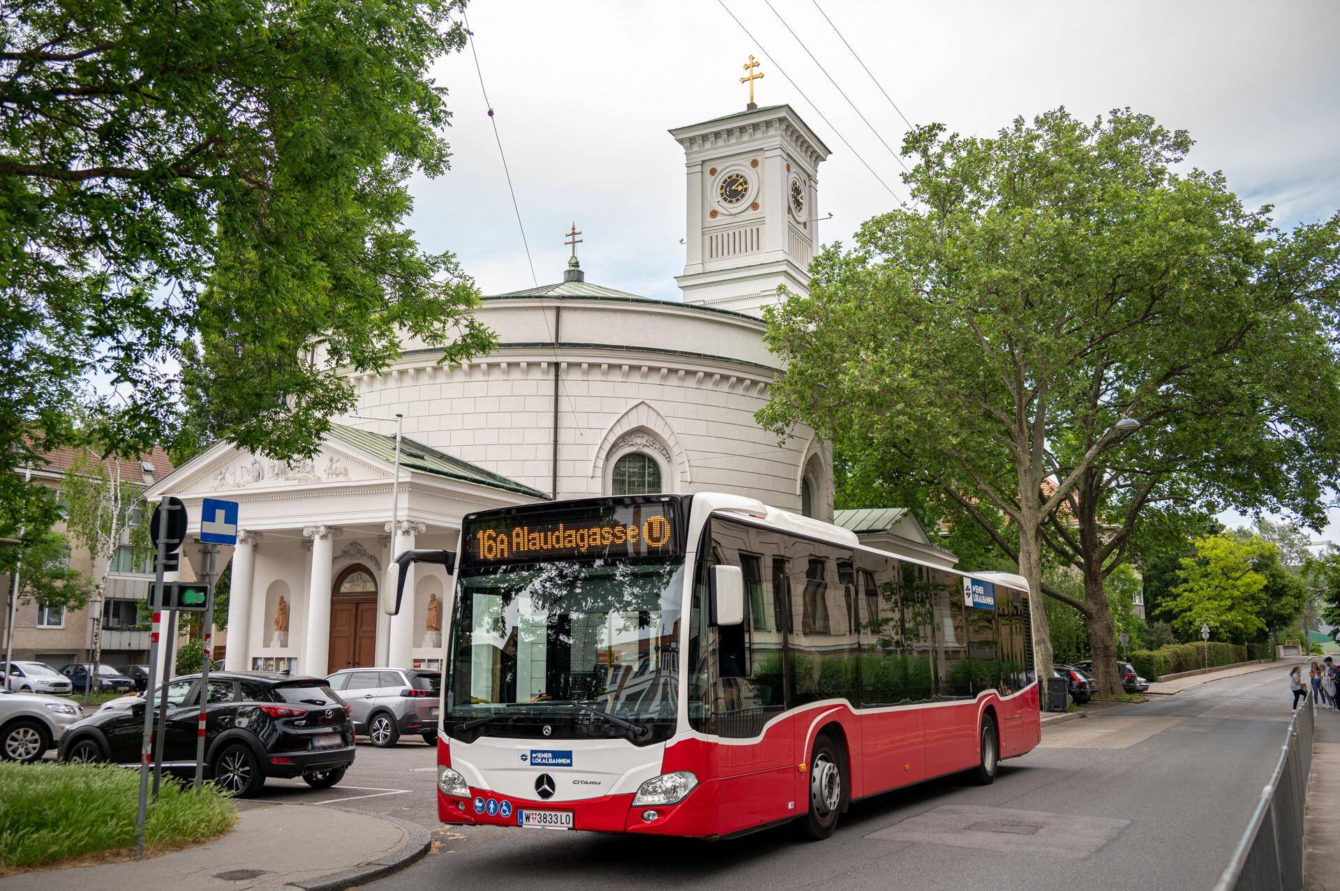 Ein moderner Bus fährt vor einer historischen Kirche mit Uhrturm in städtischer Umgebung.
