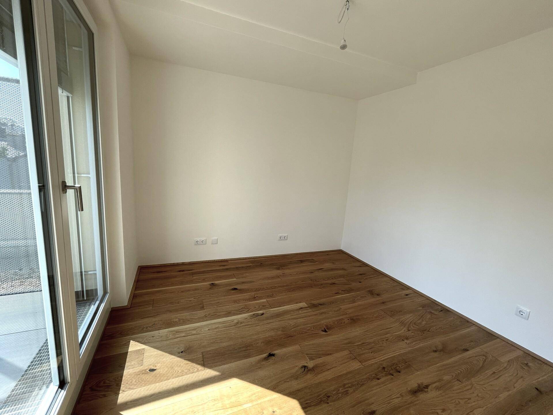 Sonniger Raum mit Holzboden und Zugang zu einem Außenbereich durch eine große Glastür.