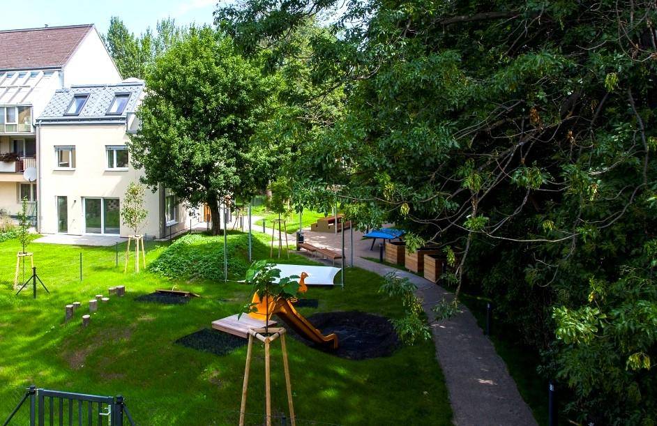 Weitläufiger Gemeinschaftsgarten mit Spielplatz, Rutsche und vielen Bäumen, umgeben von Wohngebäuden.