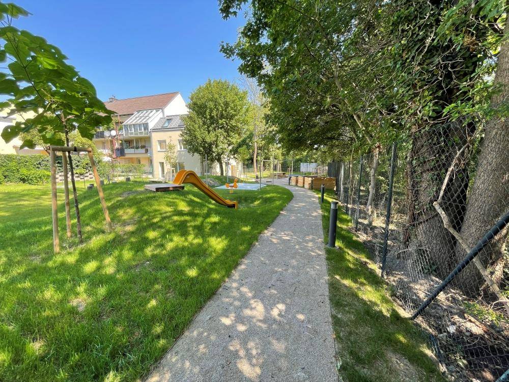 Geschwungener Weg durch den grünen Gemeinschaftsgarten mit Bäumen und Spielgeräten im Hintergrund.