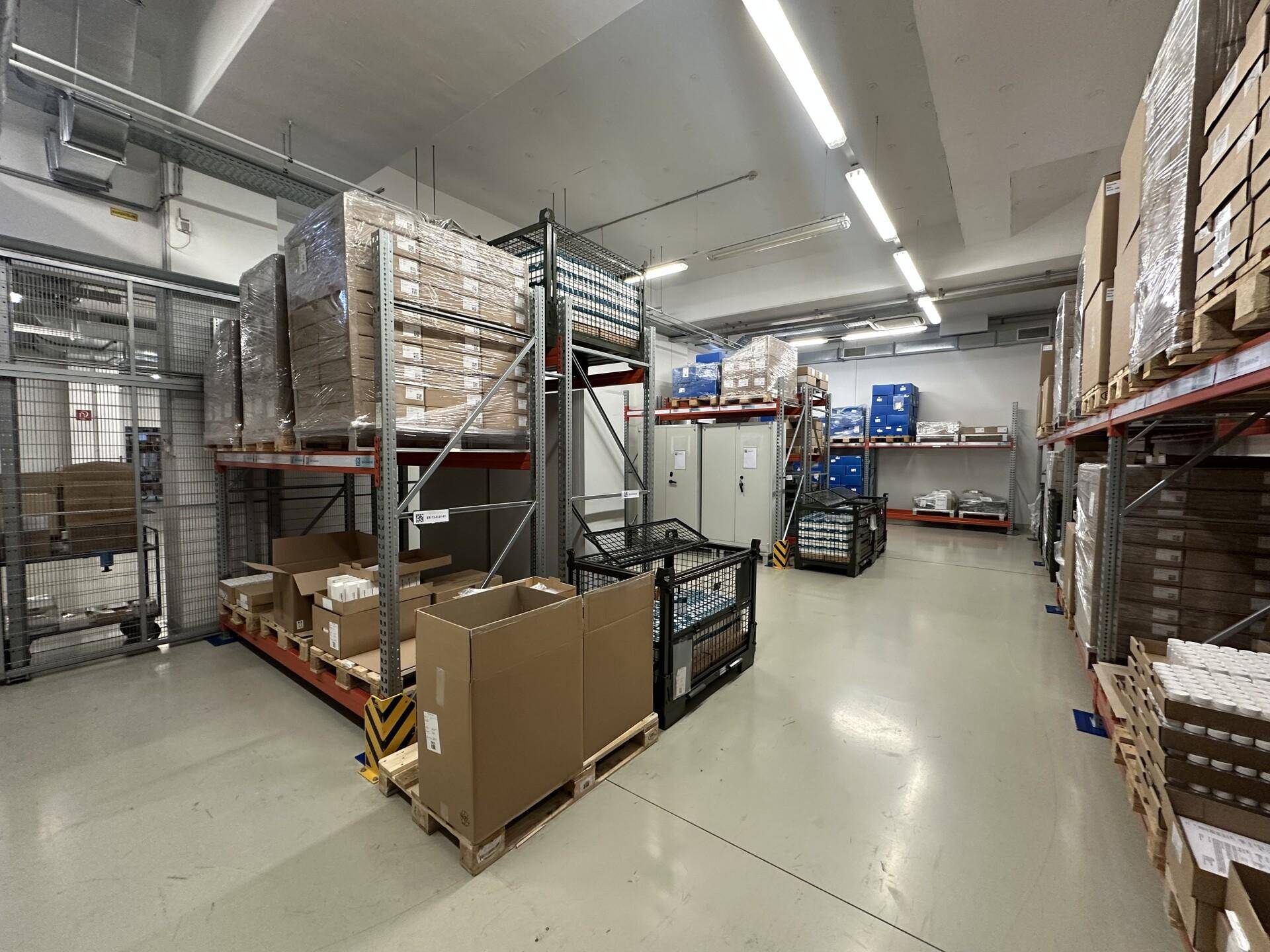 Lagerbereich mit hohen Metallregalen, gefüllt mit verpackten Gütern und einem Gitterzaun im Hintergrund.
