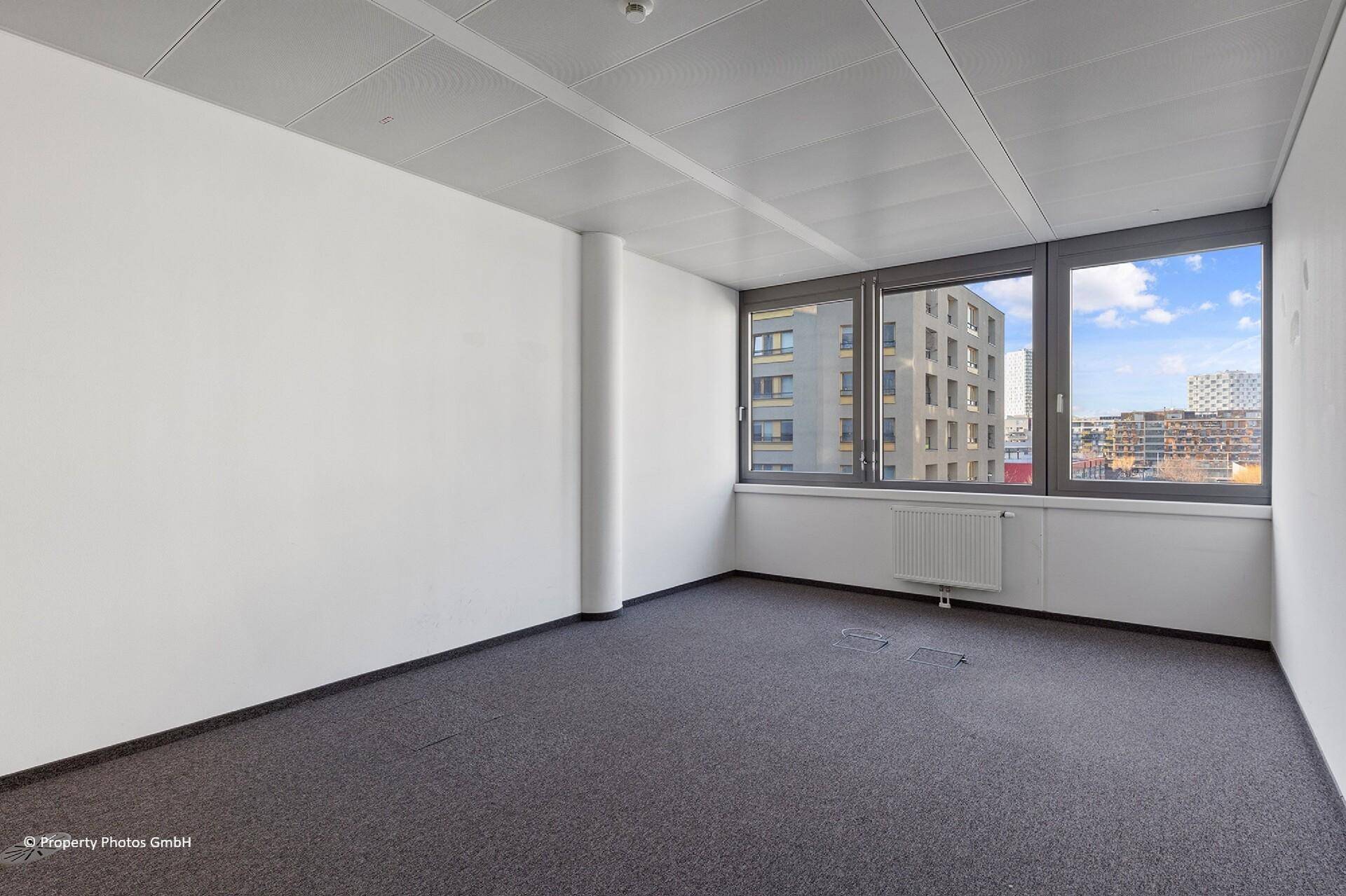 Leeres Büro mit Teppichboden, großen Fenstern und einer runden Säule, ideal für flexible Bürolösungen.
