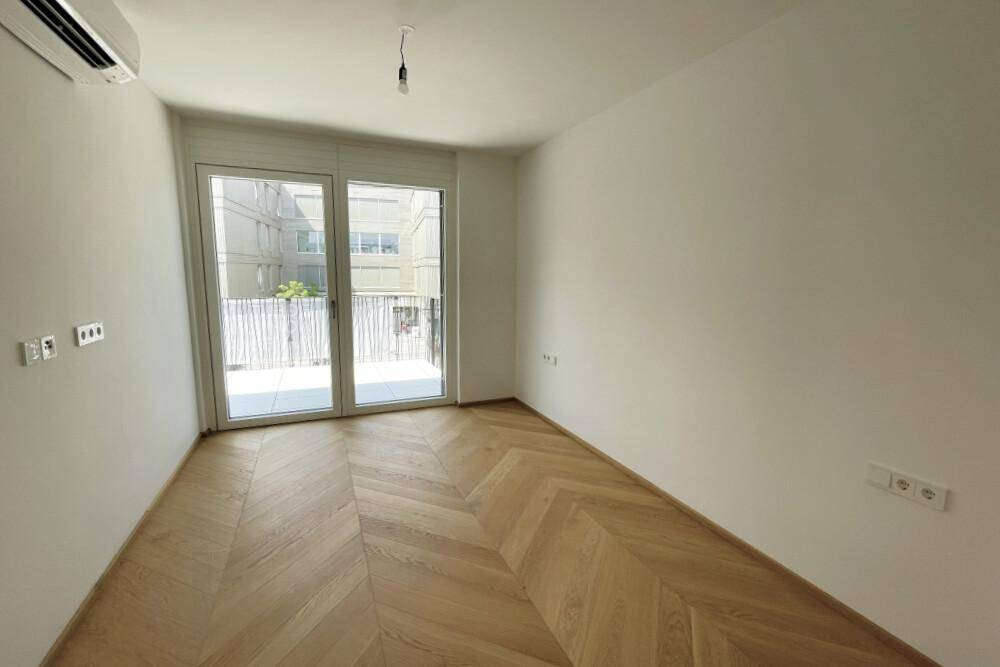 Leeres Zimmer mit Fischgrätparkett, großen Fenstern und Klimaanlage.