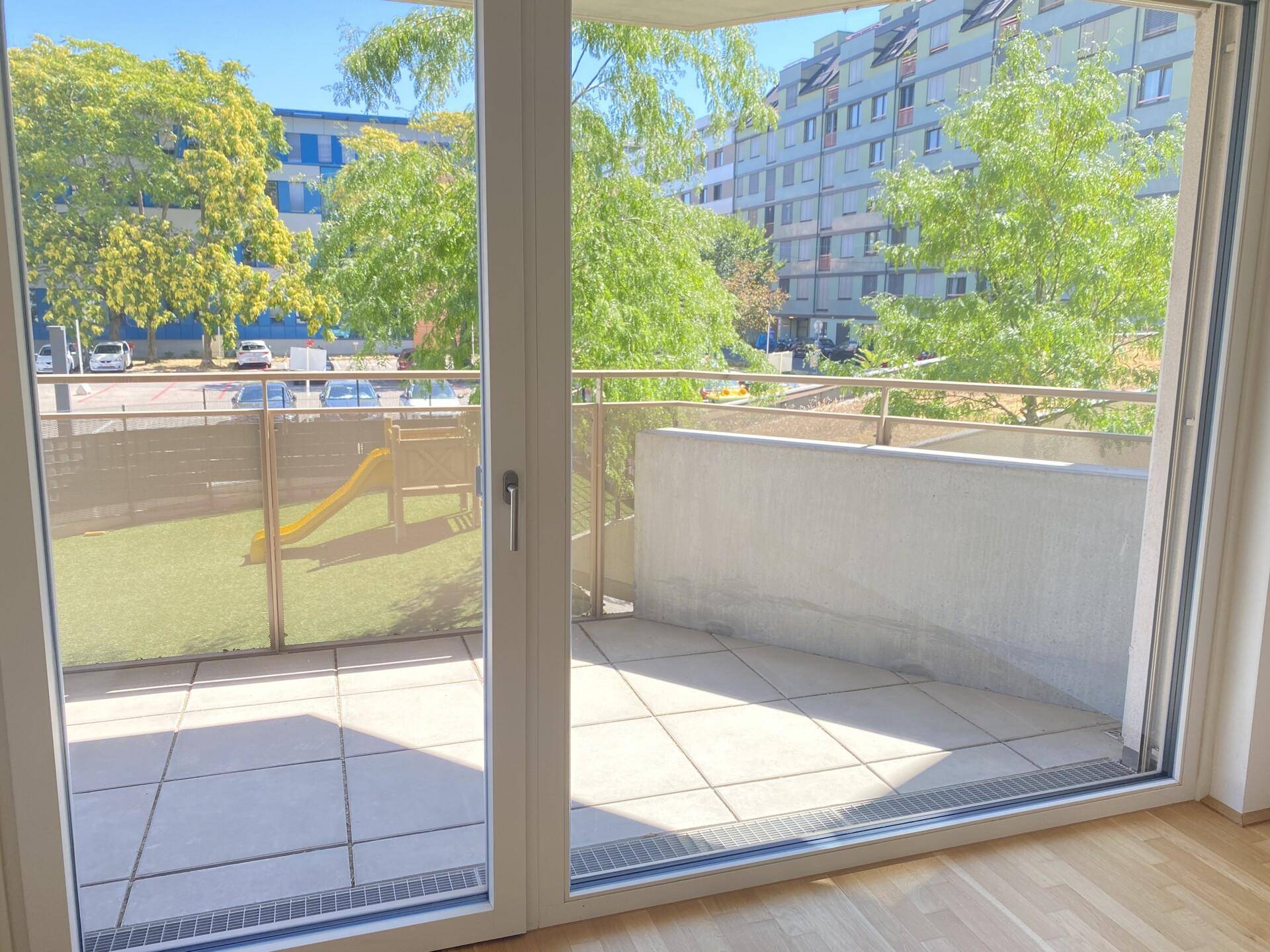 Blick aus dem Innenraum durch große Glastüren auf einen Balkon und Spielplatz.