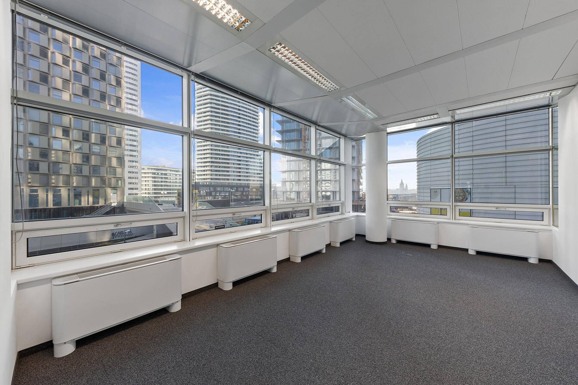 Helles Büro mit Teppichboden, großen Fenstern und Blick auf die umliegende moderne Stadtarchitektur.