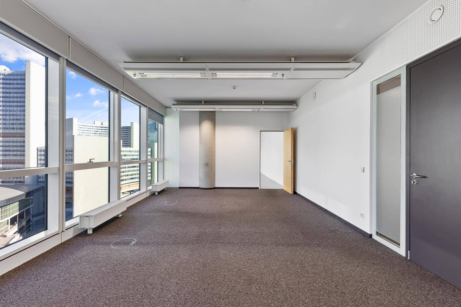 Langes, leeres Büro mit Teppichboden, großen Fenstern und zwei Türen, bereit zur individuellen Gestaltung.