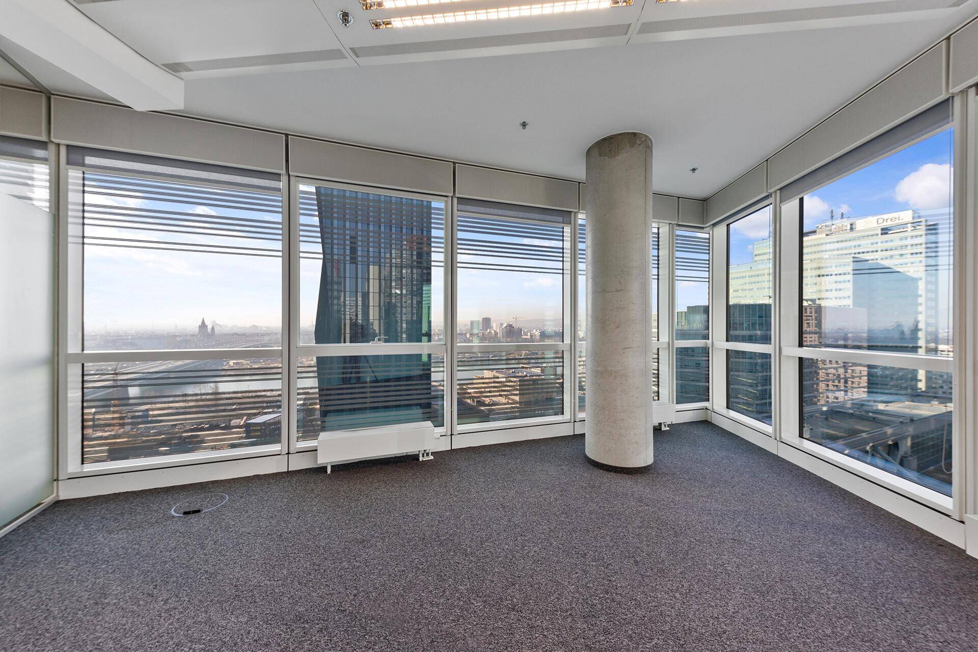 Helles Eckbüro mit Teppichboden, einer Säule und weitem Stadtblick durch die großen Fenster.