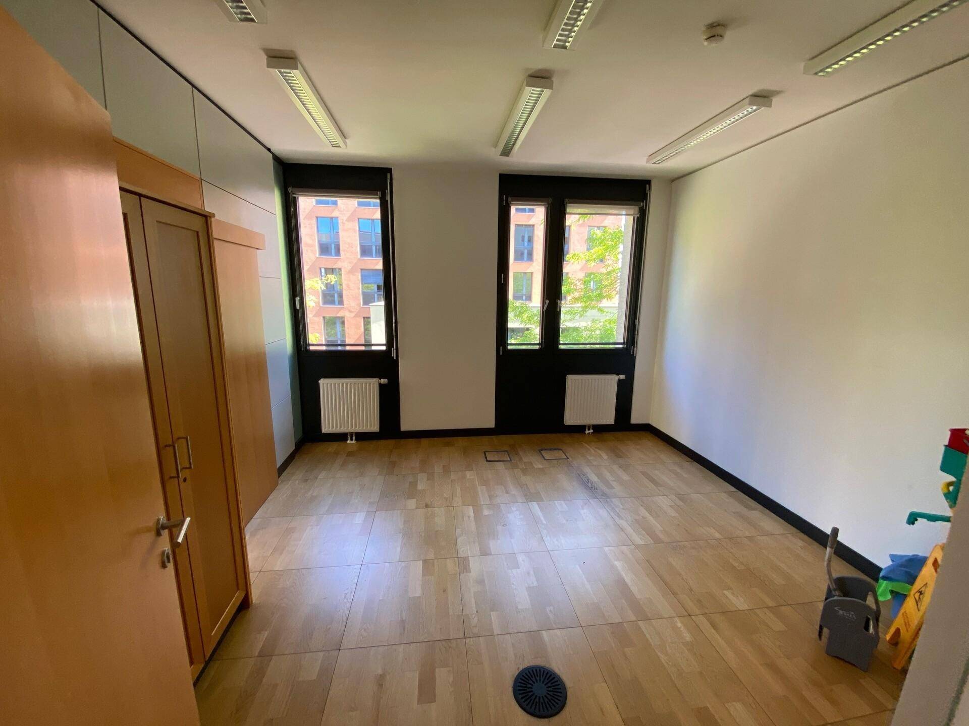 Leerer Büroraum mit Parkettboden, großen Fenstern und einem Schrank an der Wand.
