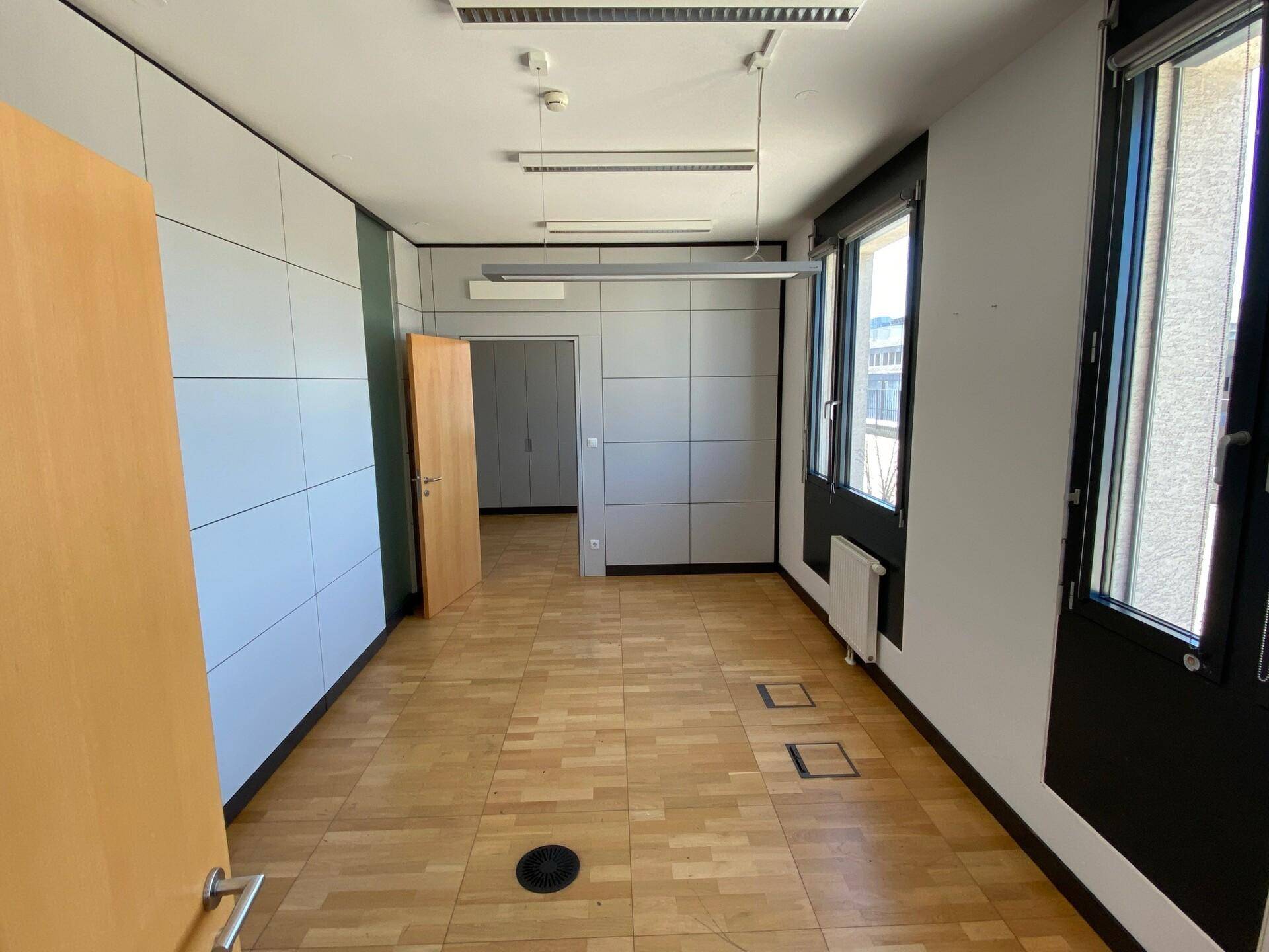 Geräumiger Büroraum mit Parkettboden und Zugang zu einem weiteren Zimmer, gut beleuchtet.