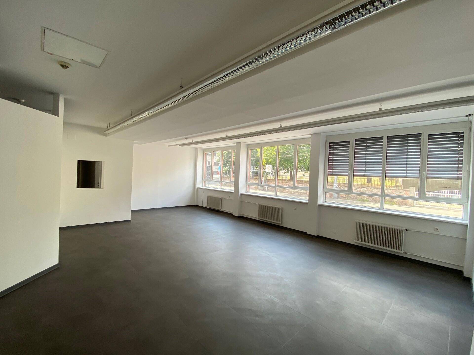 Geräumiger, leerer Büroraum mit dunklem Boden, weißen Wänden und Fenstern mit Jalousien.