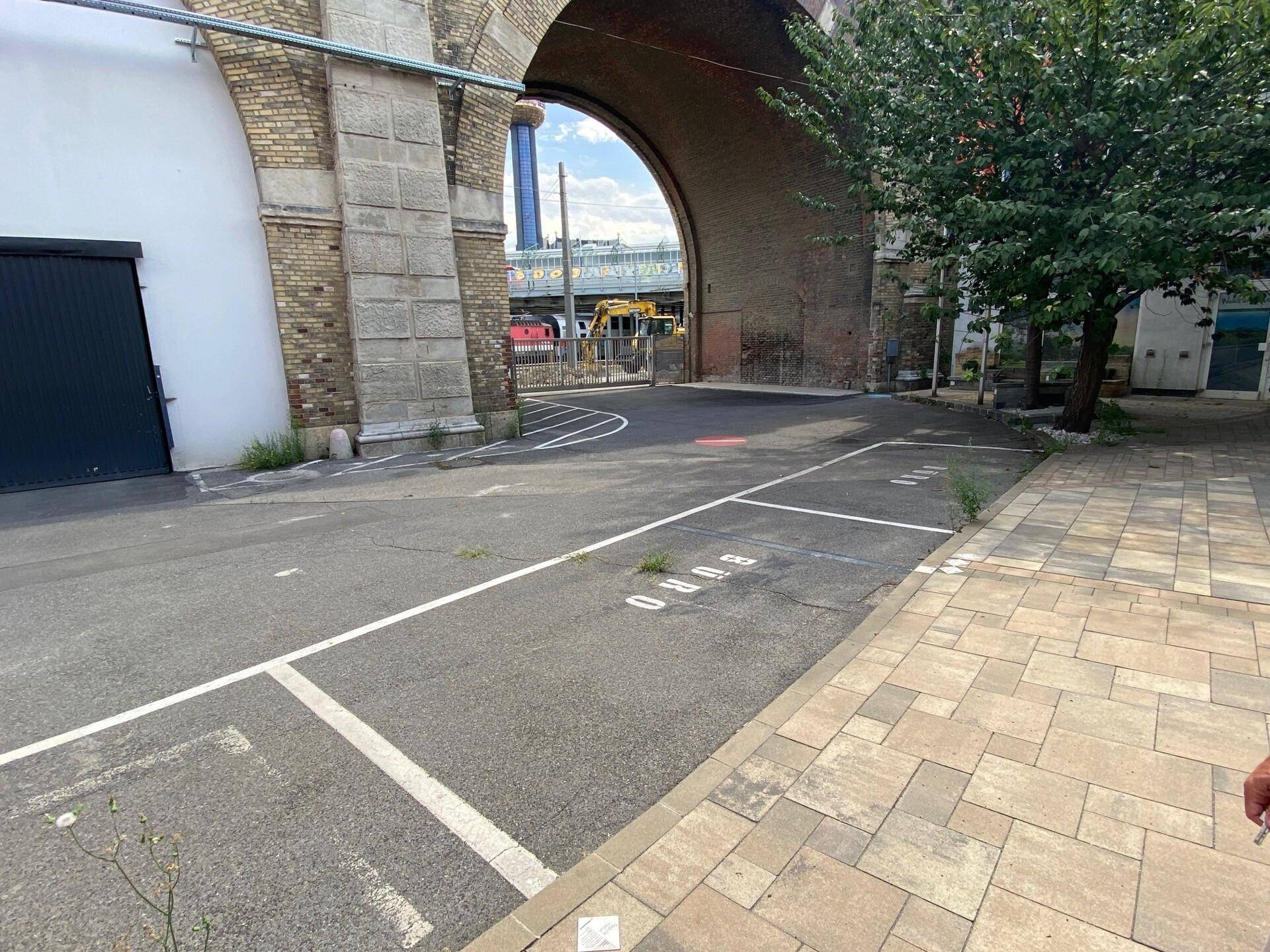 Außenparkplatz unter einer Brückenkonstruktion mit markierten Stellplätzen und angrenzendem Gehweg.