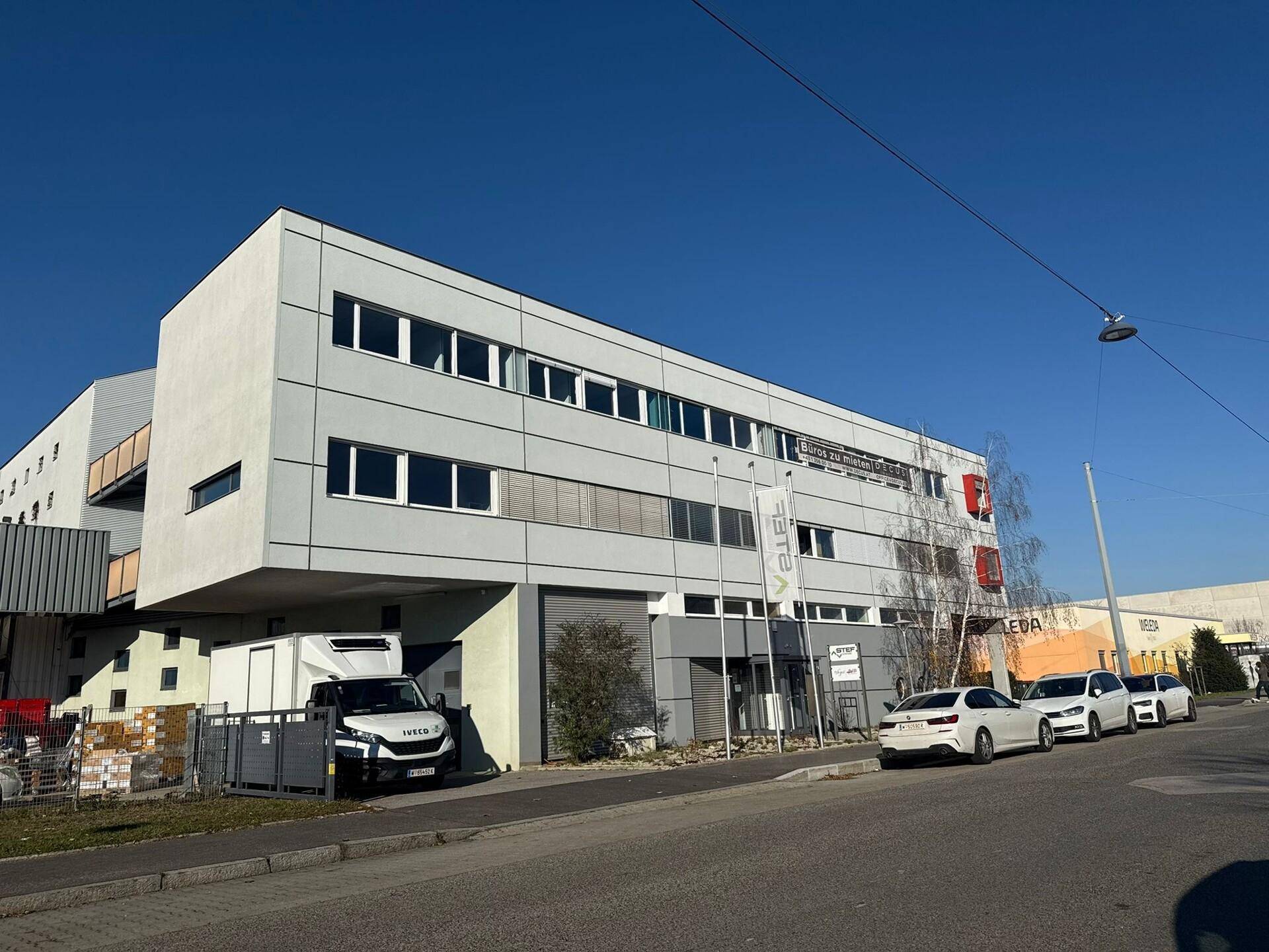 Weitläufiges Bürogebäude mit moderner Architektur und geparkten Autos auf der Straße unter blauem Himmel.