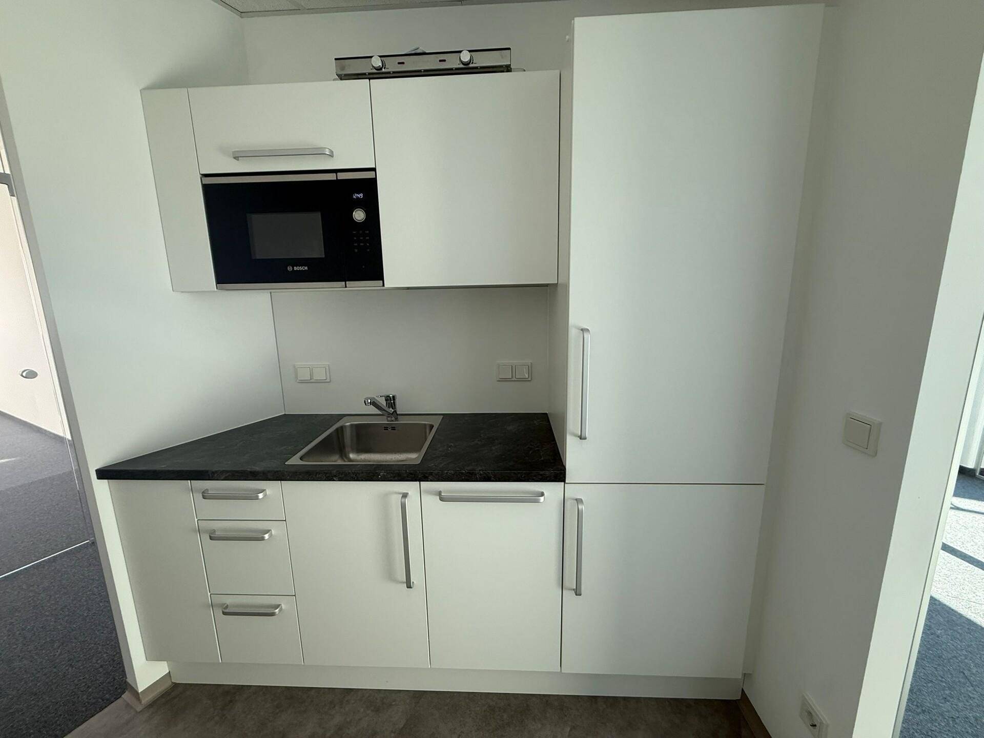 Kleine, moderne Küchenzeile mit weißen Schränken, dunkler Arbeitsplatte und integrierter Mikrowelle, in neuwertigem Zustand.