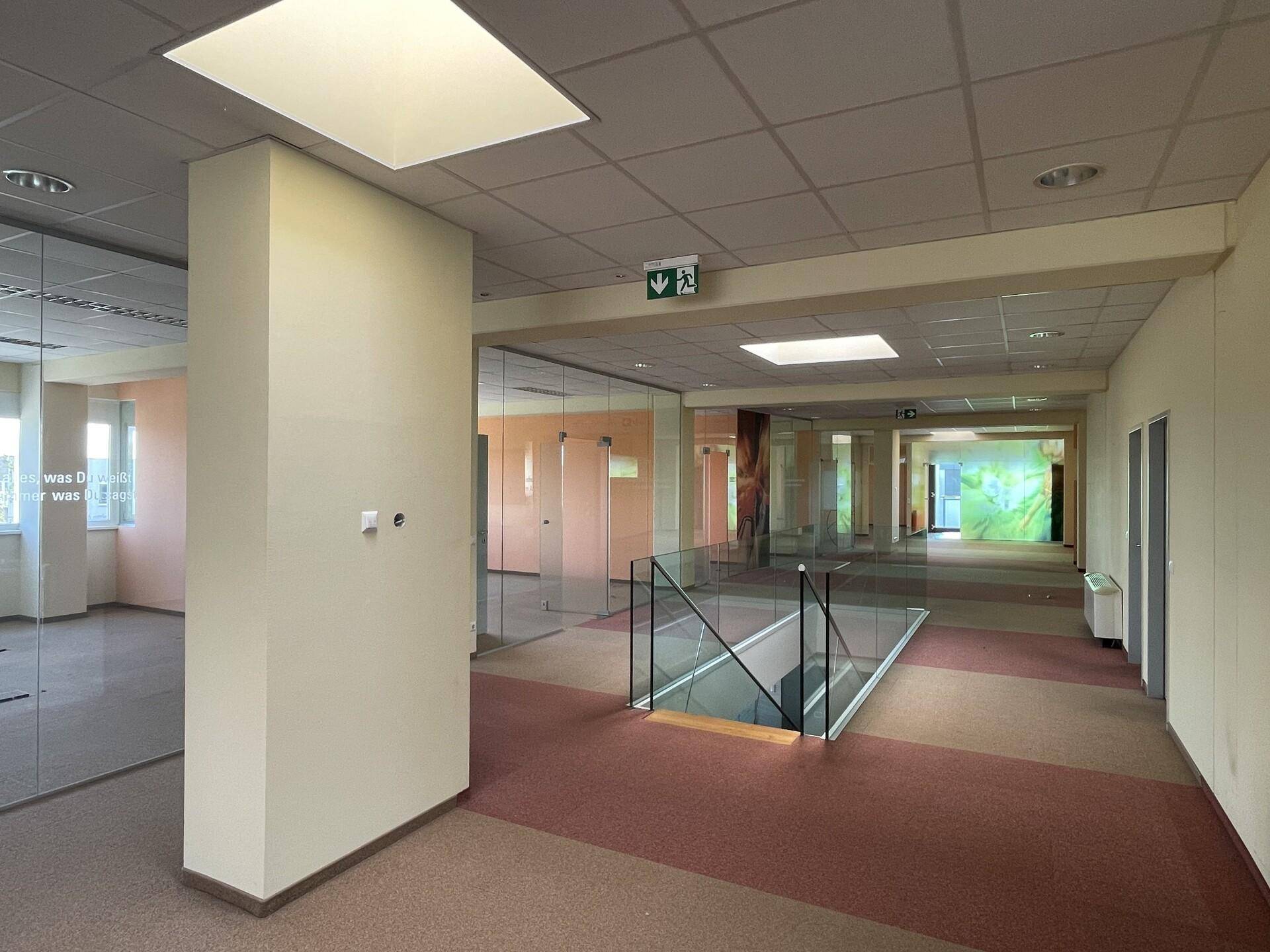 Geräumiger Flur mit Glaswänden, Teppichboden und Treppenabgang in einem Bürogebäude.