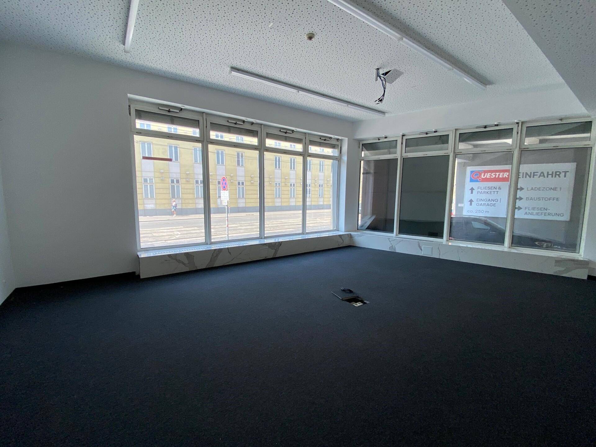 Geräumiger, leerer Büroraum mit dunklem Teppichboden und großen Fenstern mit Blick auf die Straße.