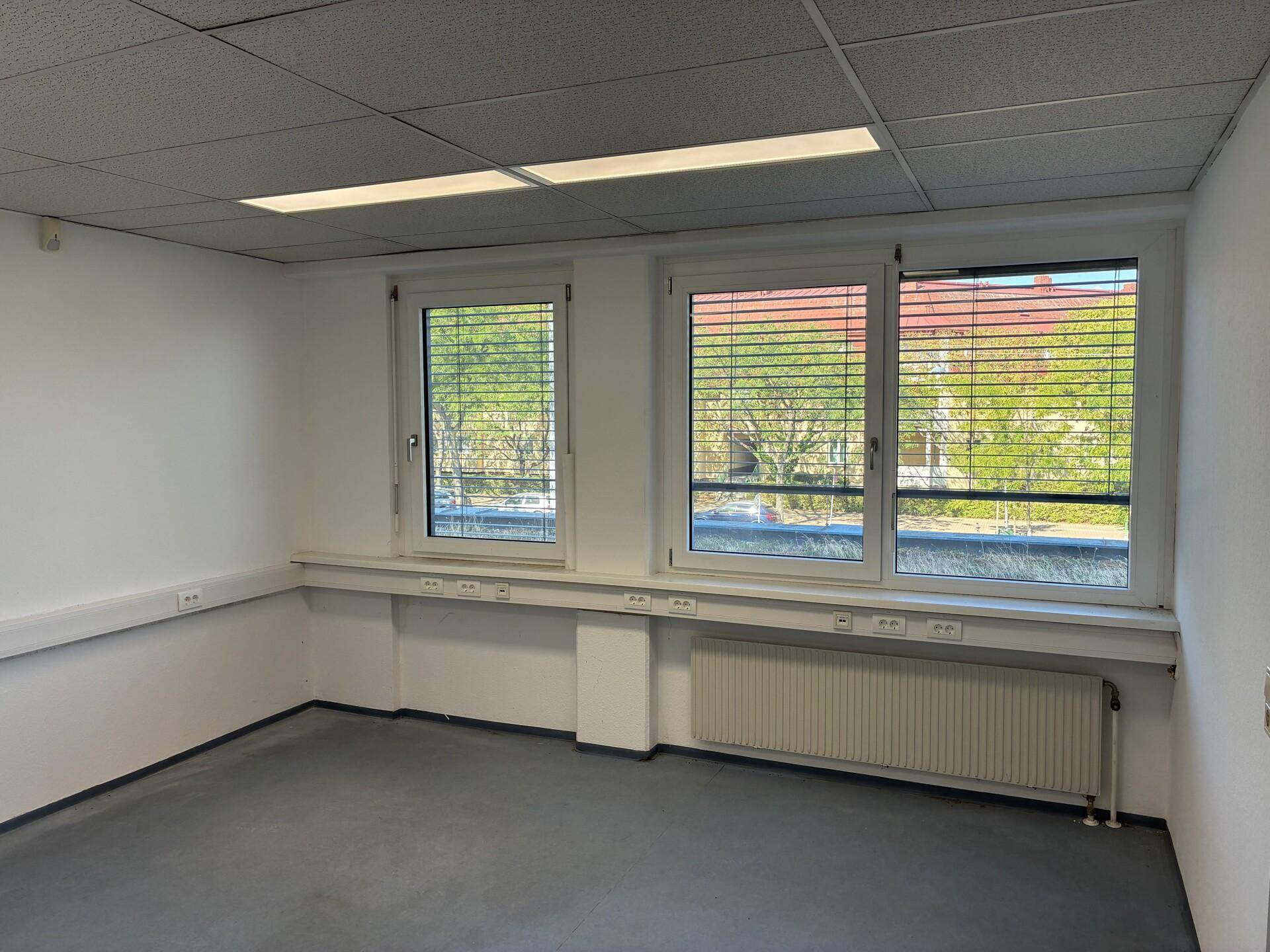 Geräumiger Büroraum mit mehreren Fenstern, Jalousien und einem Heizkörper unter der Fensterbank.