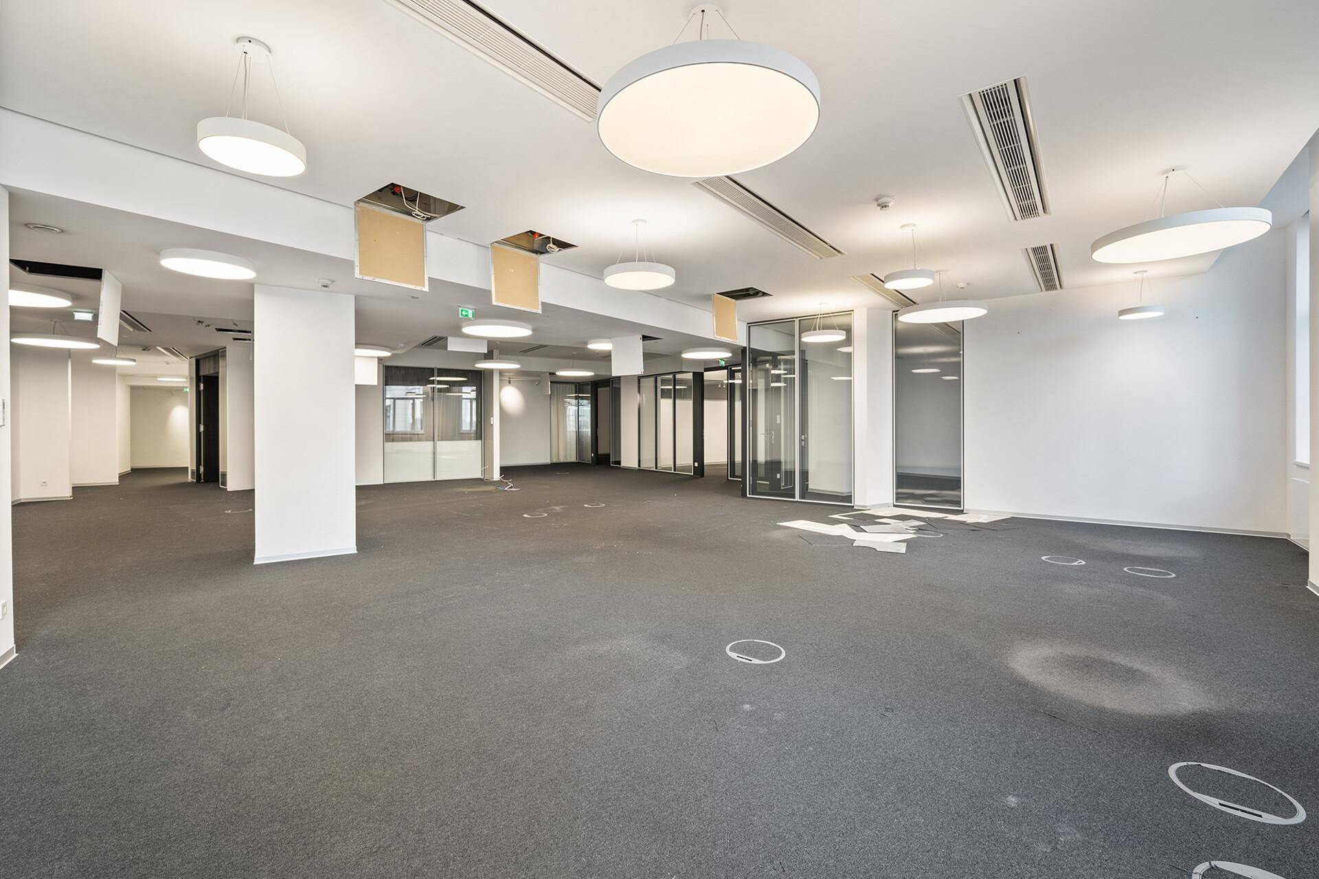 Großer, offener Bürobereich mit Teppichboden, runden Deckenleuchten und Glasabtrennungen.