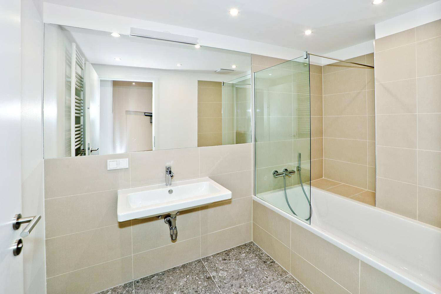 Modernes Badezimmer mit beigefarbenen Fliesen, großem Spiegel und Badewanne mit Duschabtrennung.
