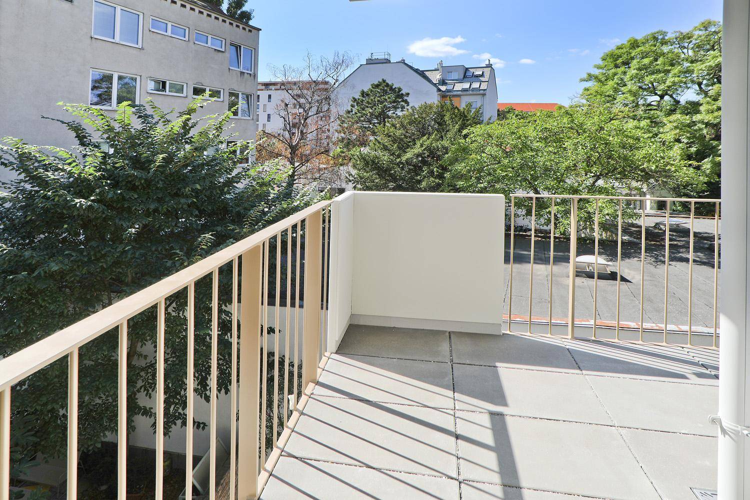 Geräumiger Balkon mit Fliesenboden und Metallgeländer, umgeben von grünen Bäumen und Gebäuden.