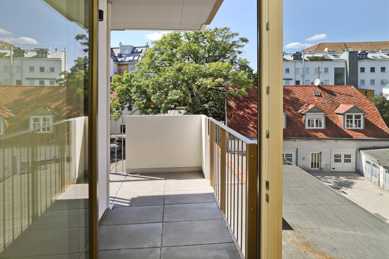 Balkon mit Fliesenboden und Blick auf die umliegenden Dächer und grüne Bäume.