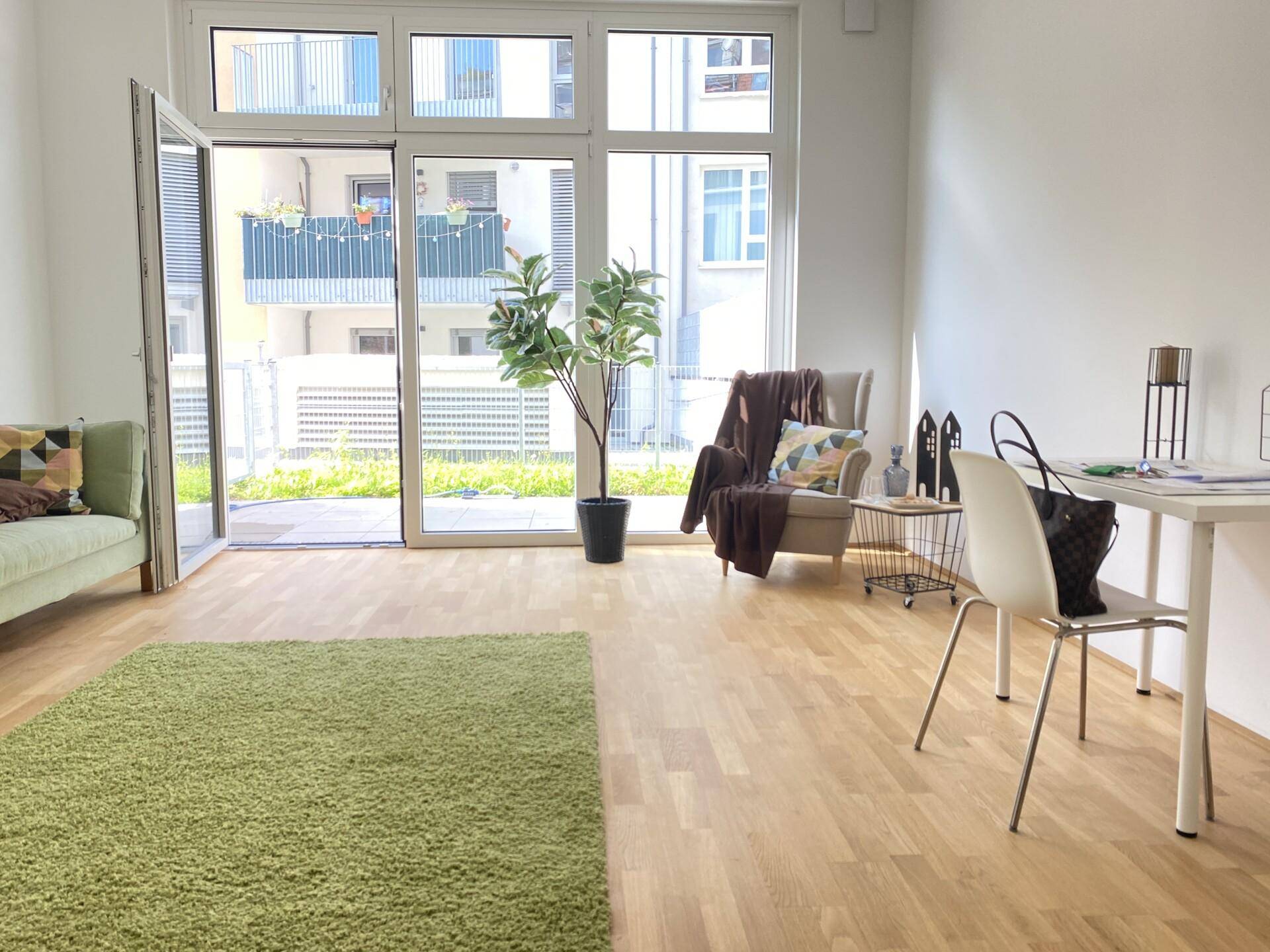 Helles Wohnzimmer mit Holzboden, grünem Teppich und großen Fenstern mit Zugang zum Außenbereich.
