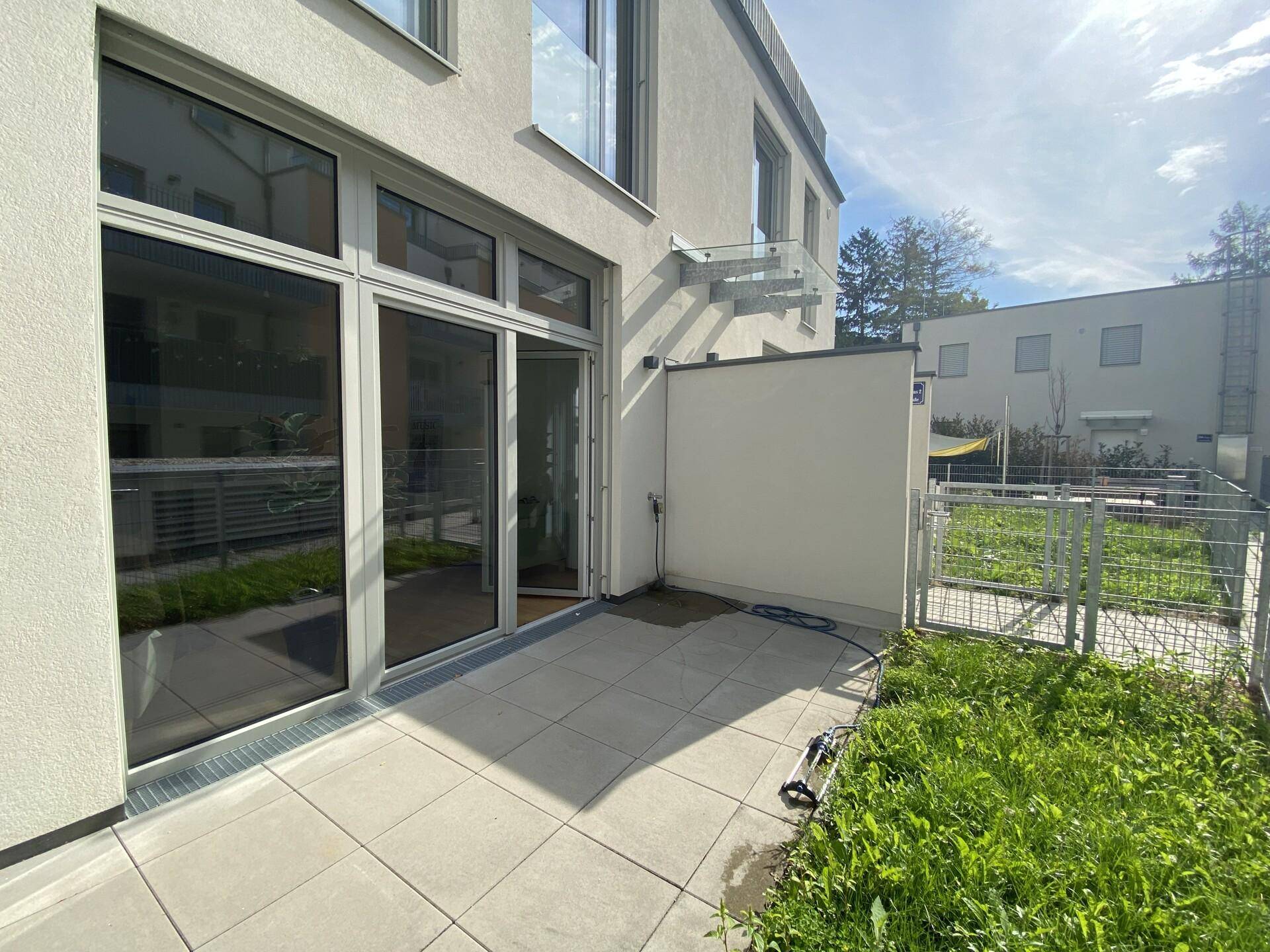 Sonniger Terrassenbereich mit Fliesenboden und angrenzendem kleinen Garten hinter einem modernen Gebäude.