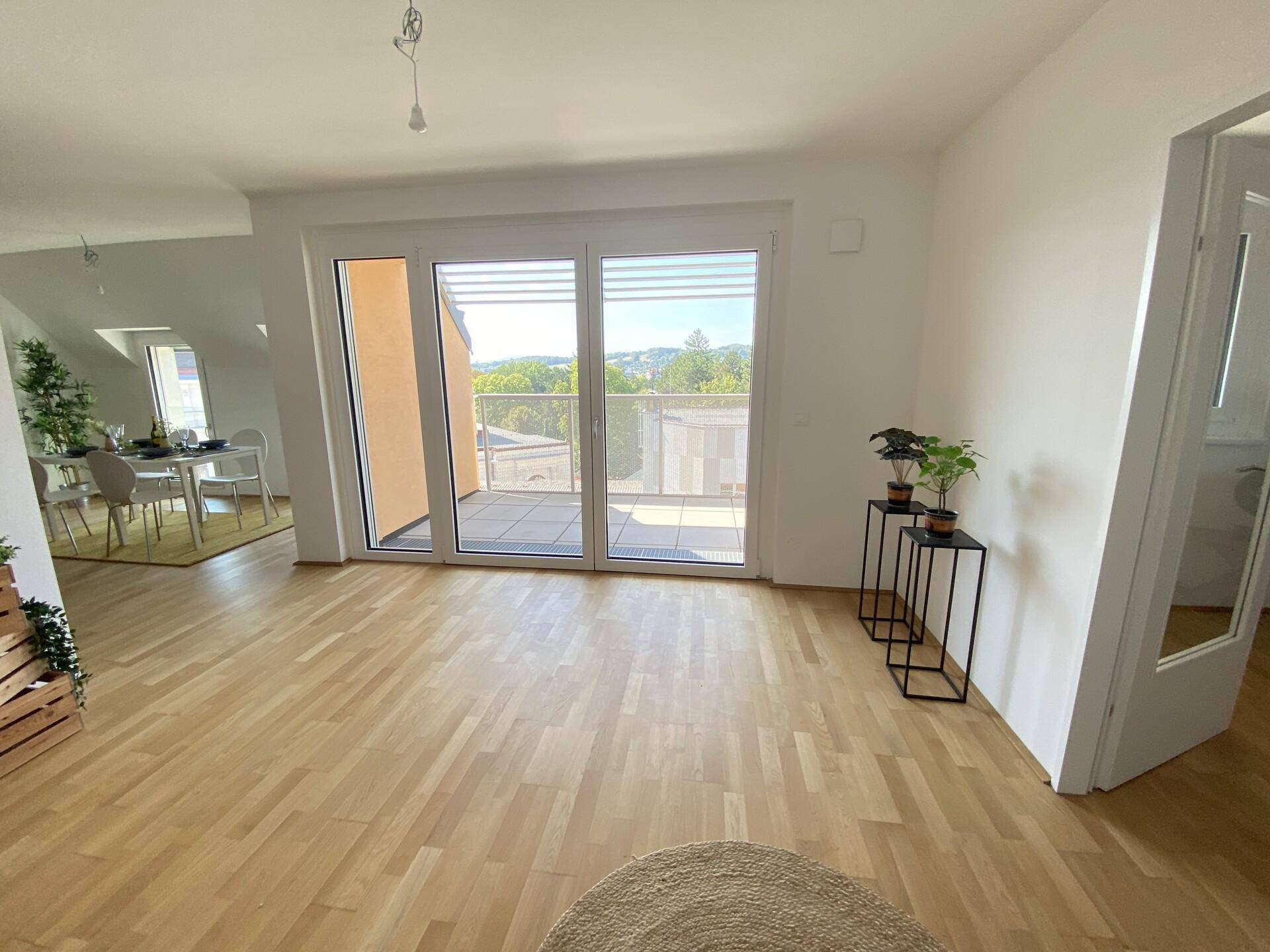 Geräumiger Wohnbereich mit Holzboden und großen Fenstern, die auf eine sonnige Terrasse führen.