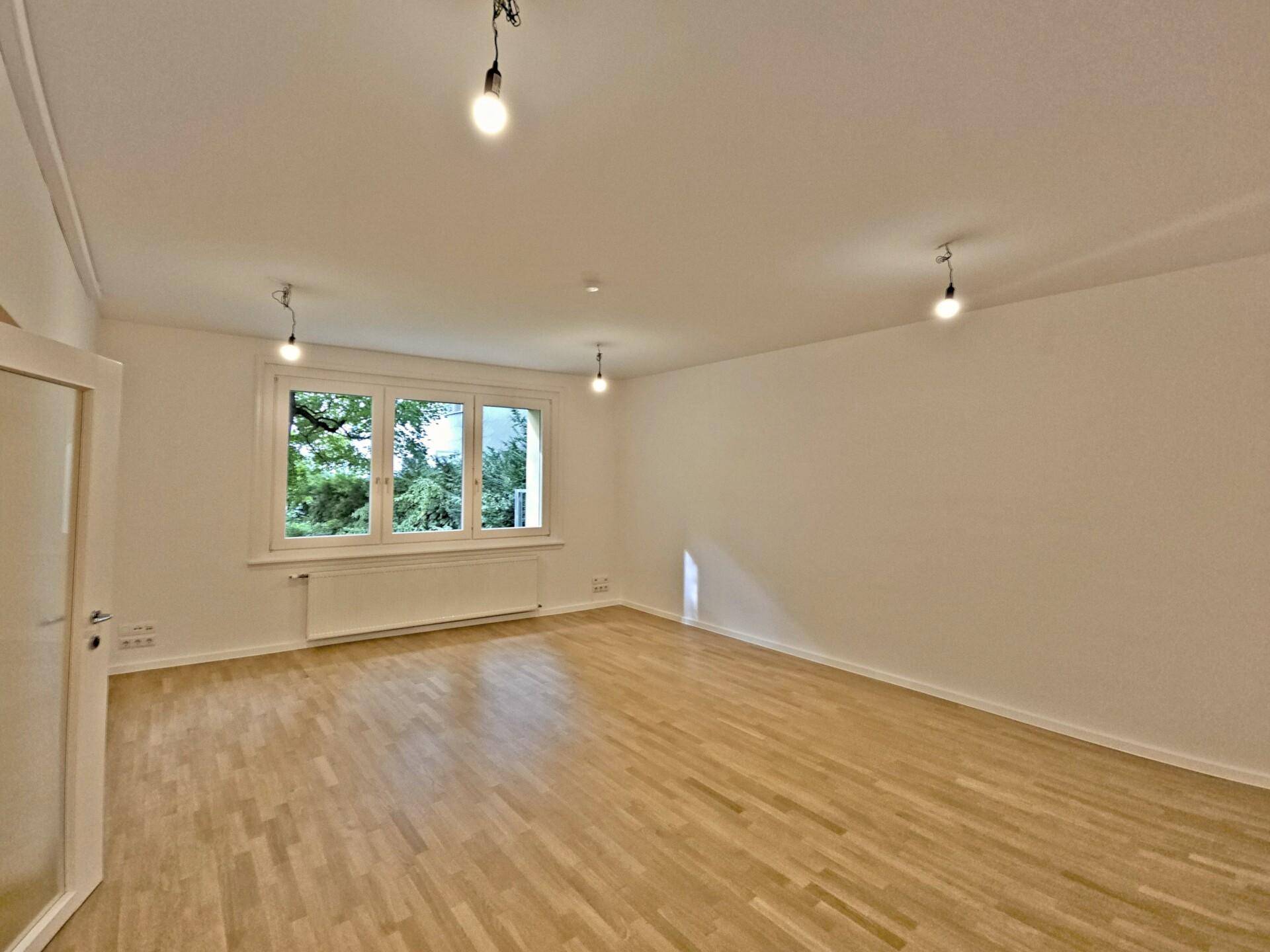 Heller, leerer Wohnraum mit Parkettboden, weißen Wänden und einem großen Fenster mit Blick ins Grüne.