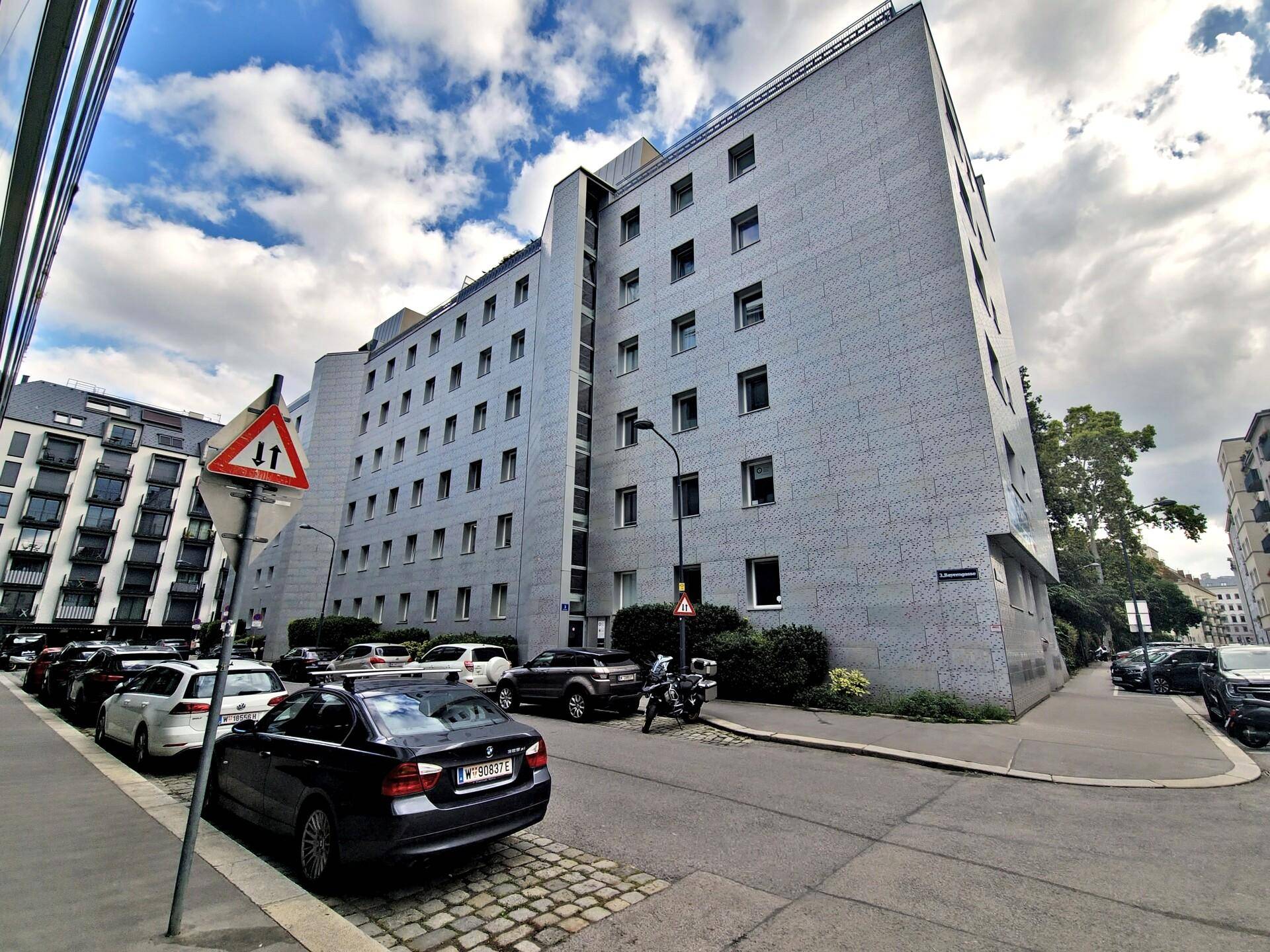 Großes, modernes Apartmentgebäude mit grauer Fassade, vielen Fenstern und parkenden Autos auf der Straße.