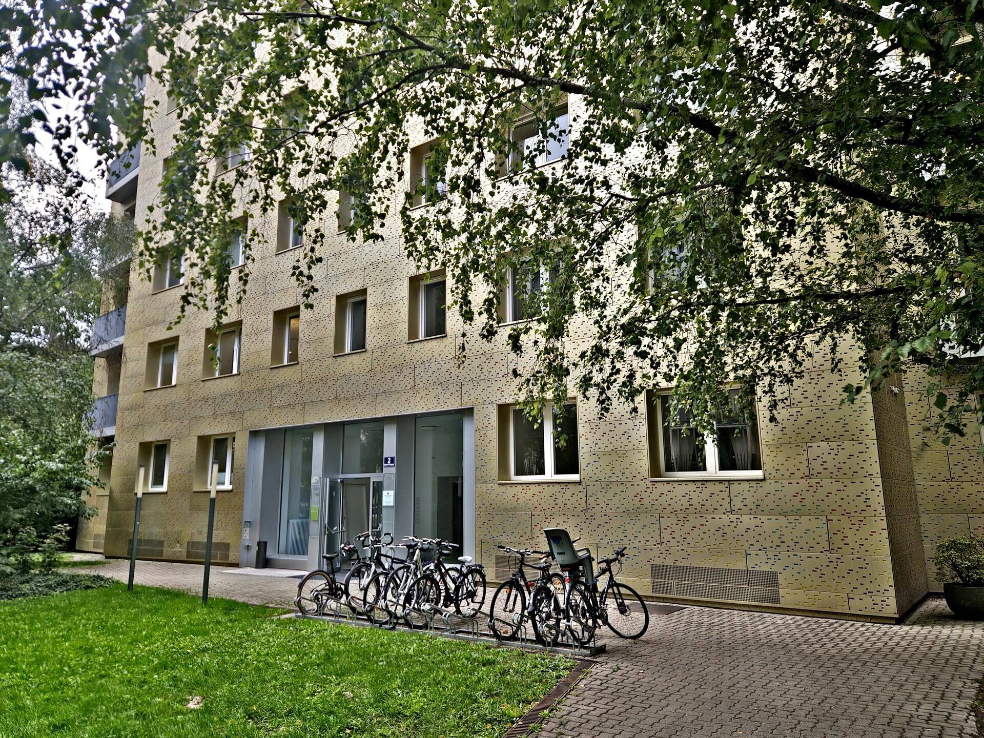 Eingangsbereich eines modernen Apartmentgebäudes mit beiger Fassade, Fahrradständern und Grünflächen.