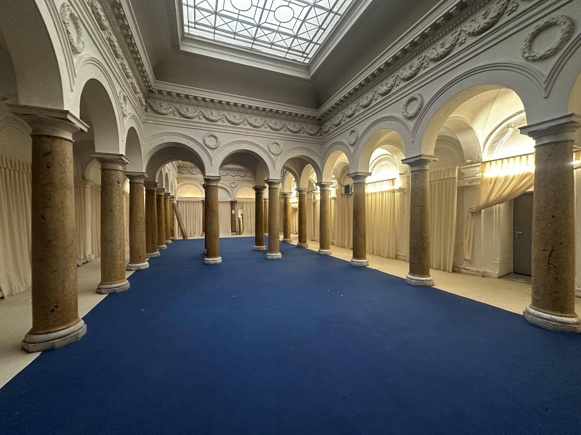 Prunkvoller, großer Saal mit Säulen, Bögen, einem Oberlicht und blauem Teppichboden, ideal für Veranstaltungen.