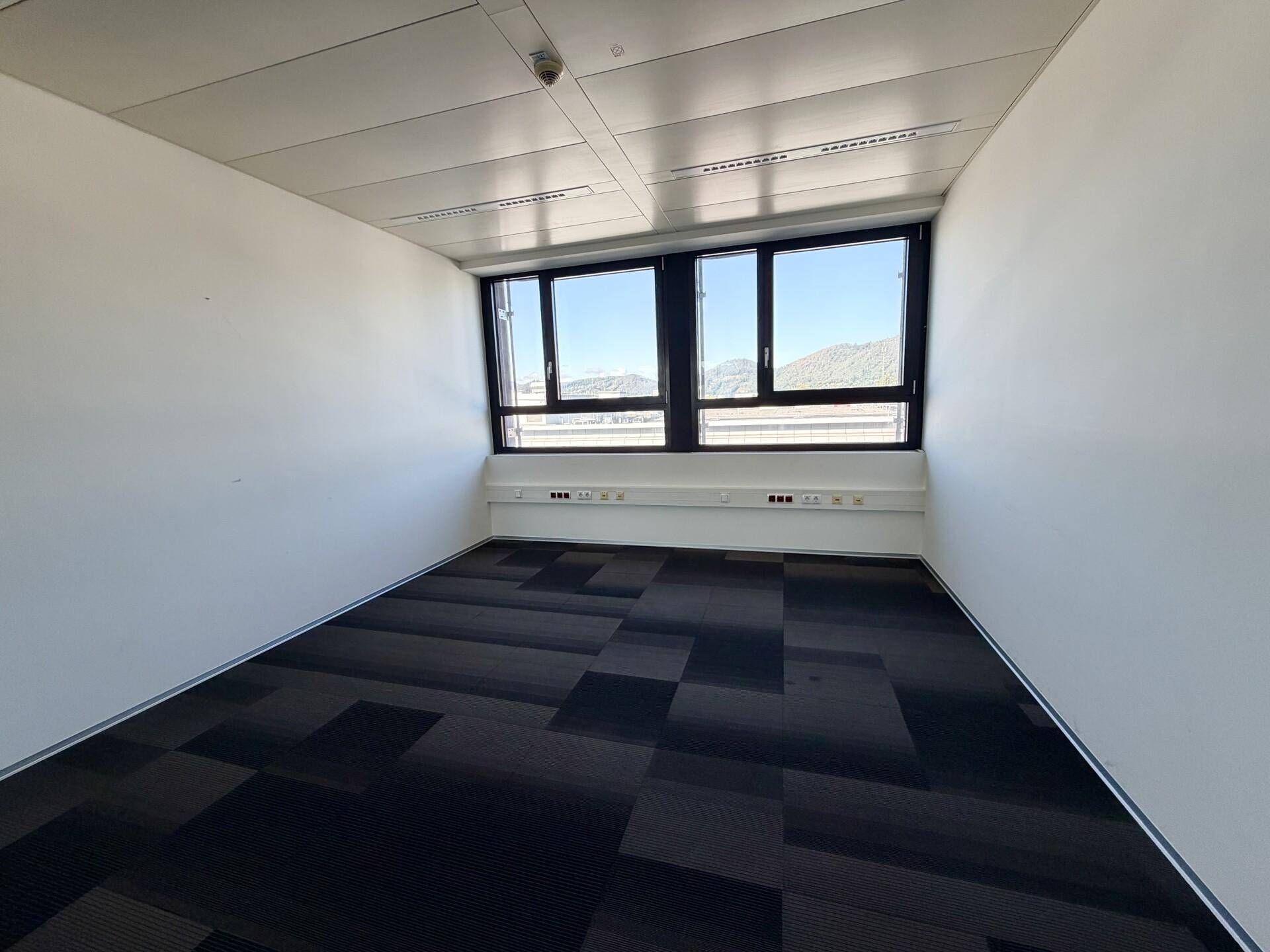Leeres Büro mit Teppichboden und großen Fenstern, die viel Tageslicht hereinlassen.