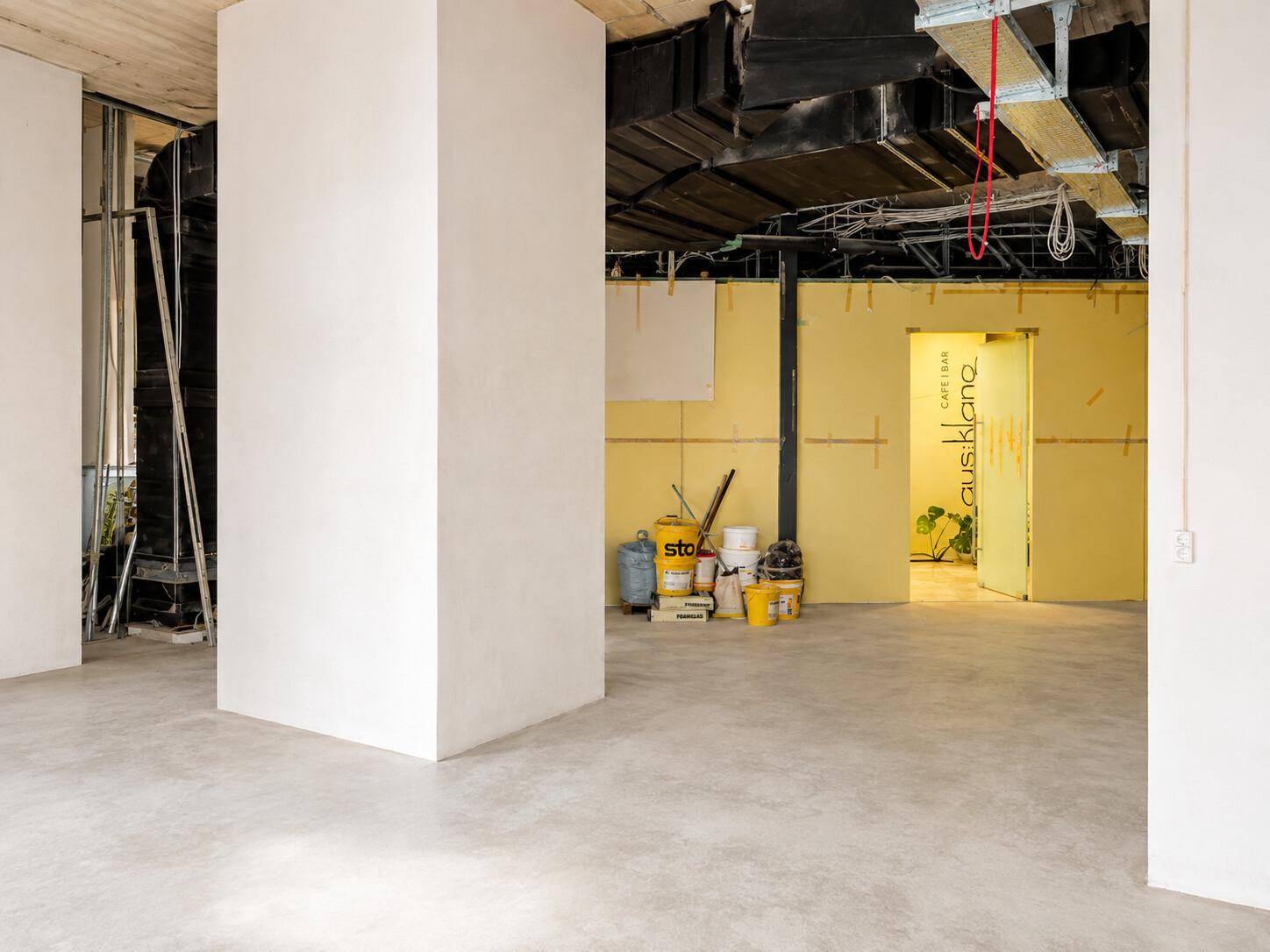 Großer, unfertiger Gewerberaum mit hohen Decken, sichtbaren Installationen und Betonboden.