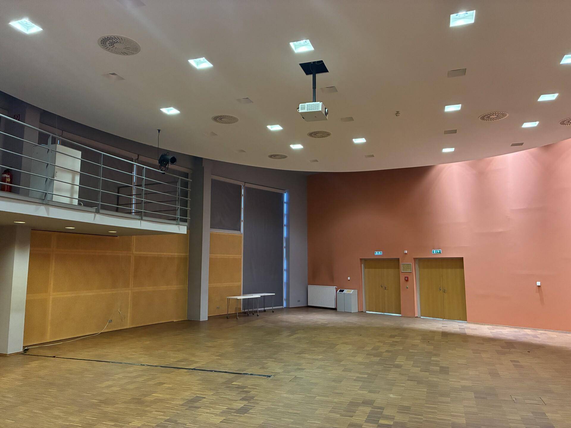 Großer Veranstaltungsraum mit Holzboden, Projektor und mehreren Ausgangstüren, ideal für Versammlungen.